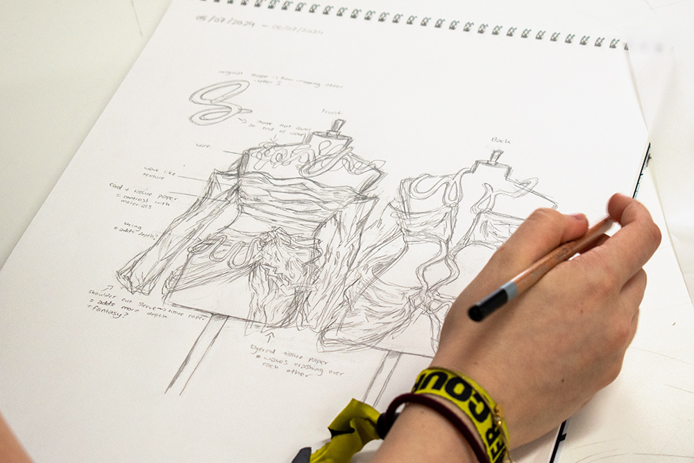 Hand holding pencil sketching mountain landscape on spiral-bound paper. Yellow and black wristband visible on artist's wrist.