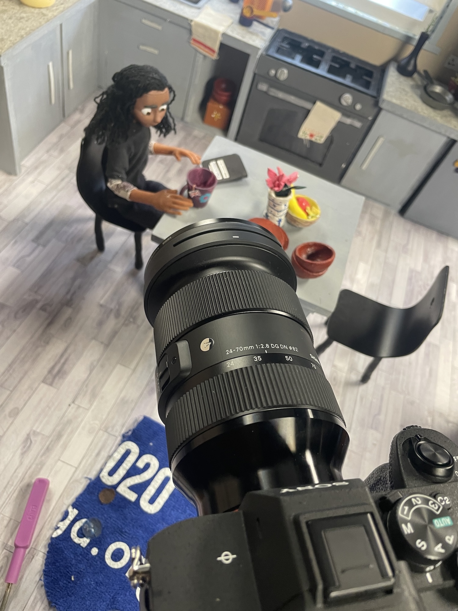 Close-up of a camera looking over an animated character sitting at a table in a kitchen. The chair opposite the character at the table is empty.