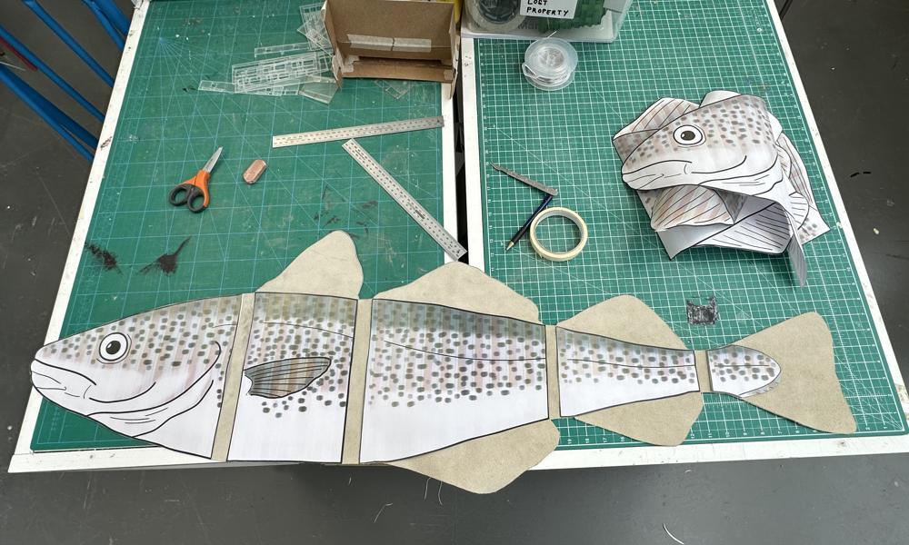 Paper cutout design of a cod separated into four sections, scissors, tape, rules and scraps of other paper parts laid out next to it on a green drawing board surface.