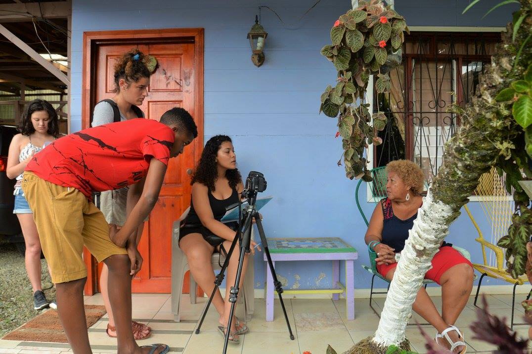 Four students filming an interview with a small camera on a tripod with an older woman, sitting on a bench outside a house on the right.