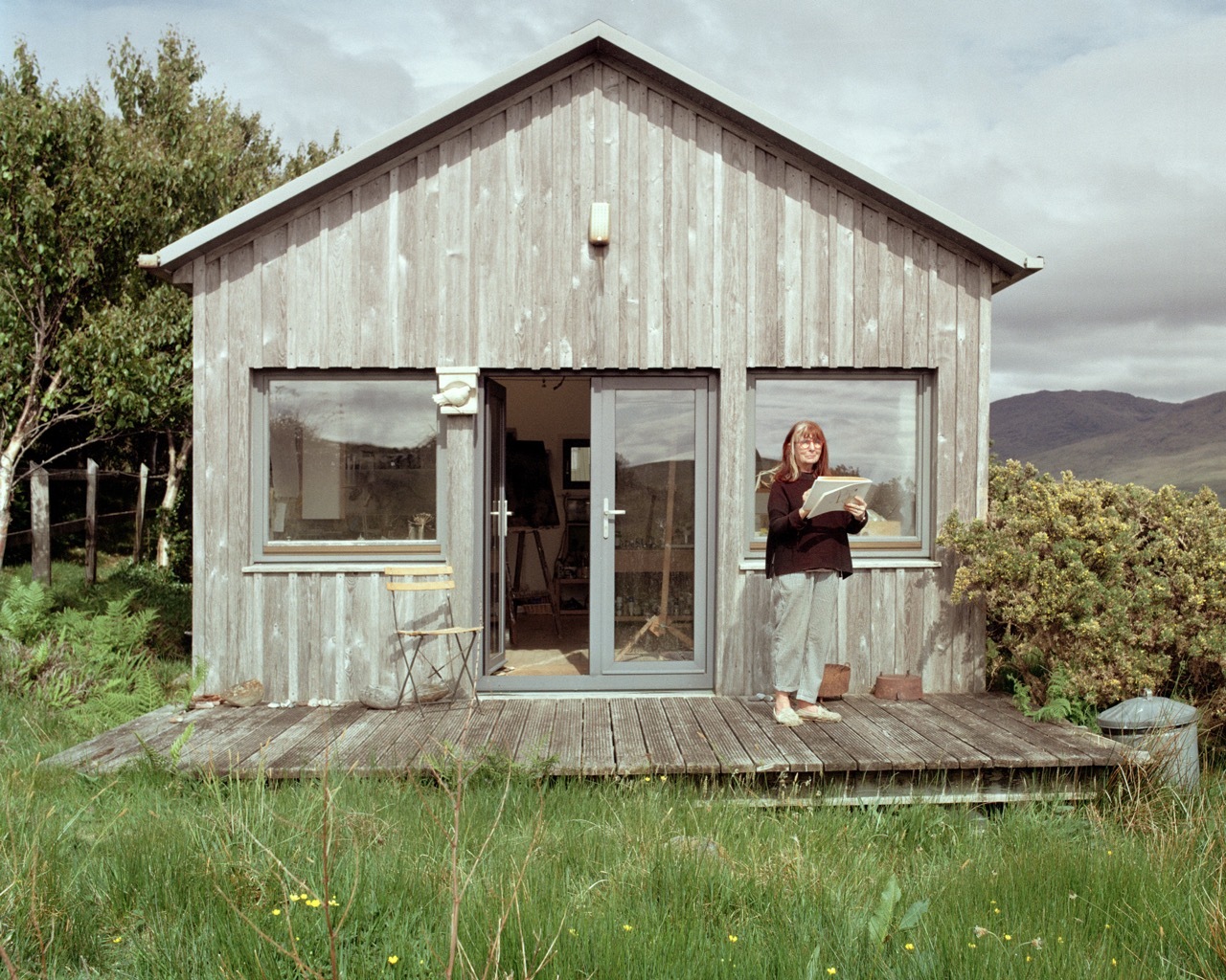 A woman stood outside a modern pale wood cabin in the wilderness, reading a book.
