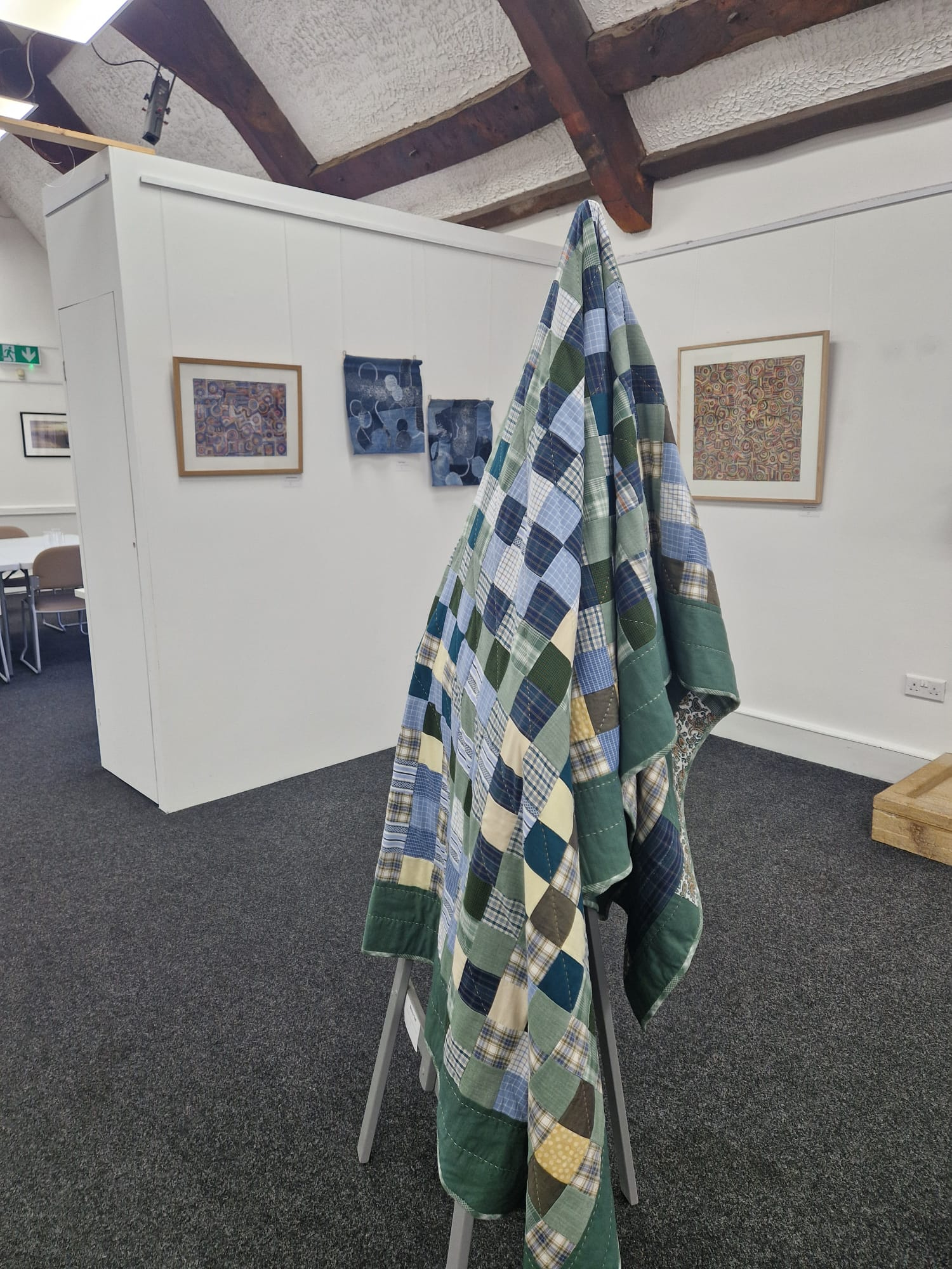 A patchwork quilt with a green, blue, and white checkered pattern draped over a metal display stand in an art gallery. In the background, framed artworks hang on the white walls, and the floor is covered with dark carpet.