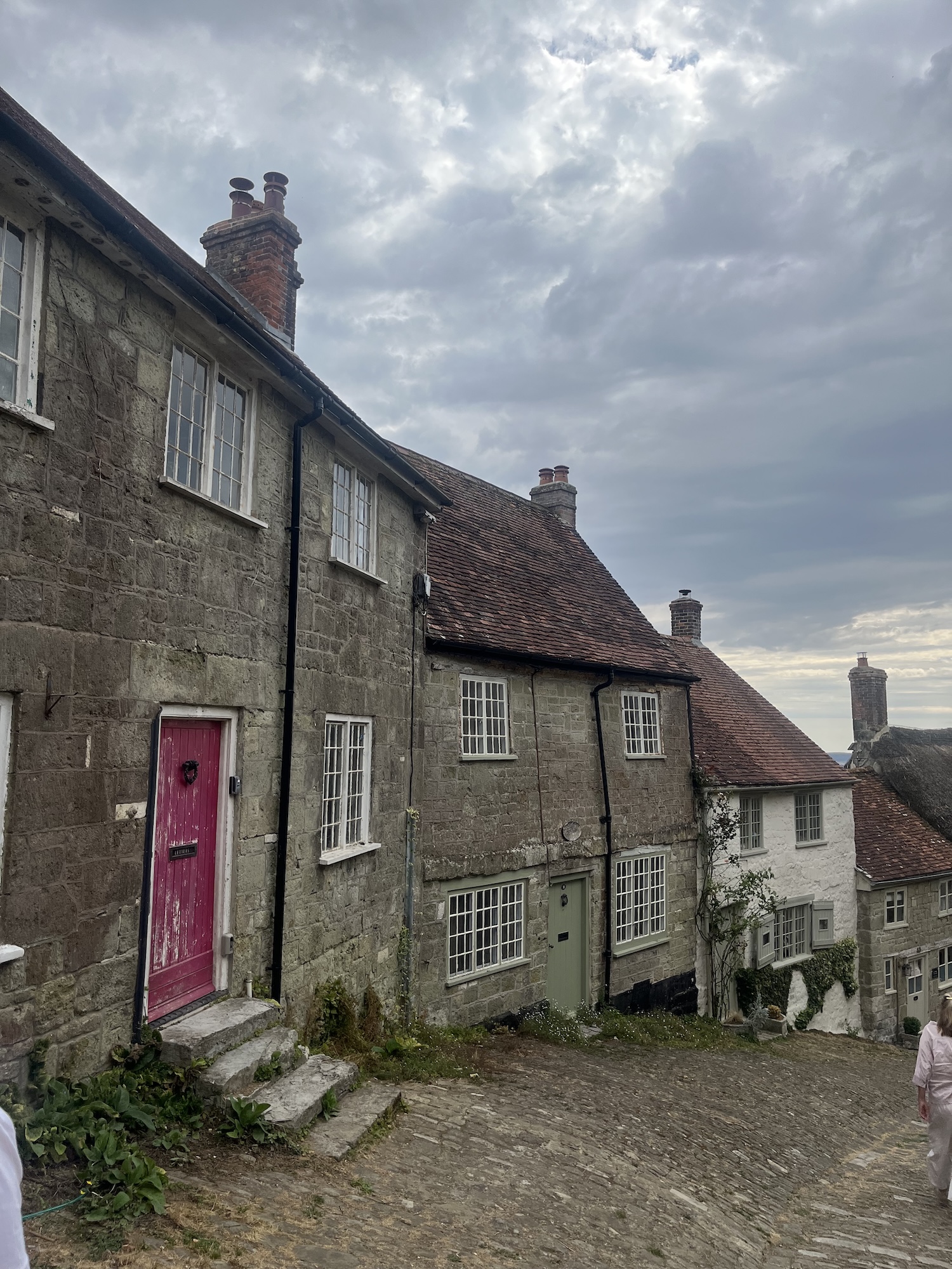 A row of stone cottages with coloured doors lines a steep cobbled street, with a cloudy sky overhead.