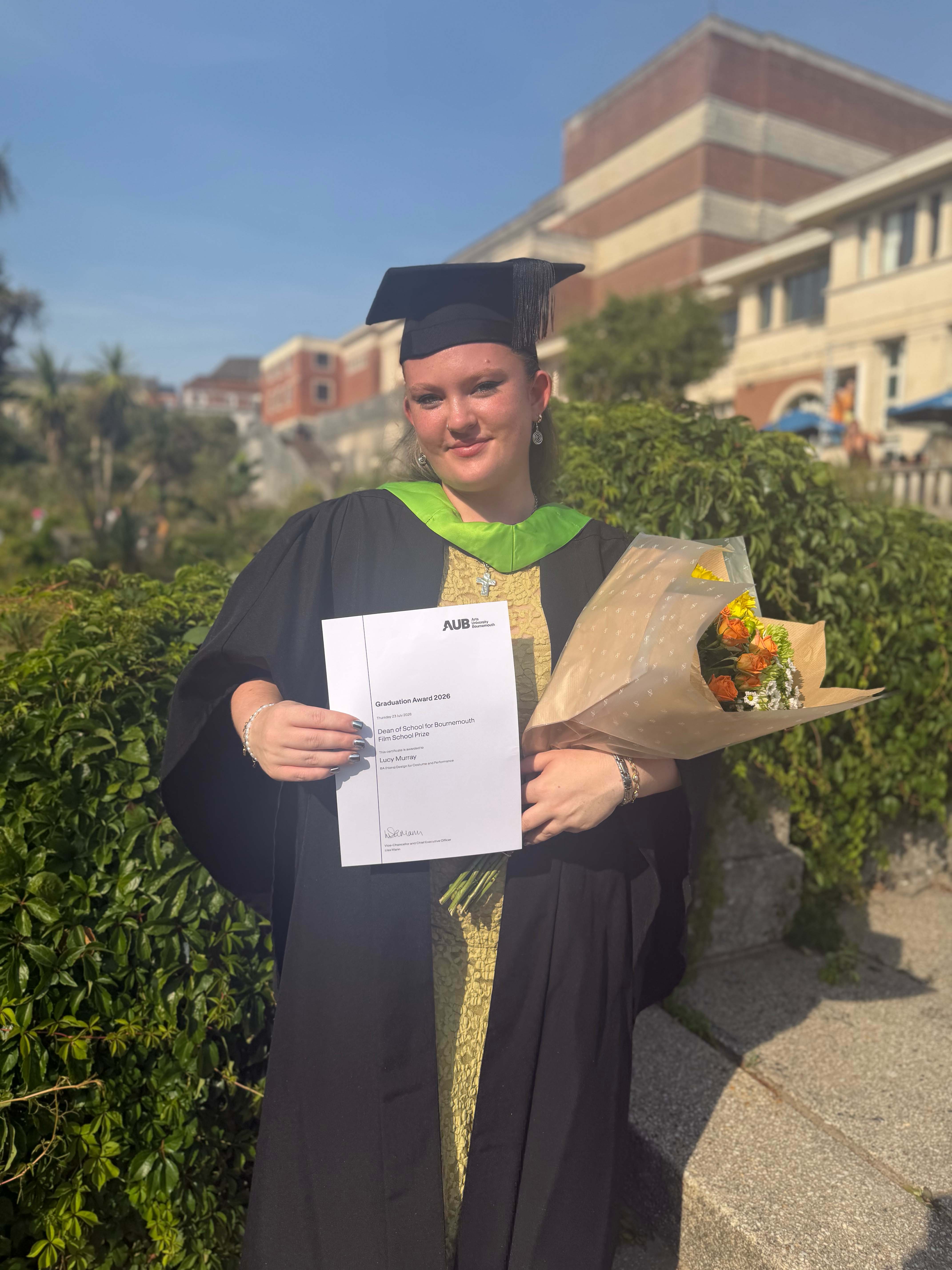 A graduate in a black gown and mortarboard holds a certificate and a bouquet of flowers outdoors, with a university building and greenery in the background.