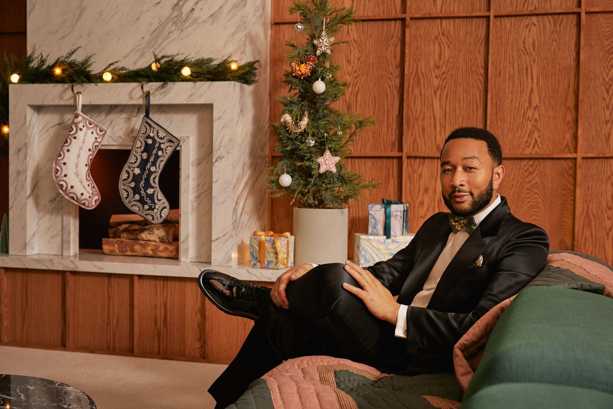 Musician John Legend sitting cross-legged on a sofa, with a fireplace and Christmas decorations in the background.