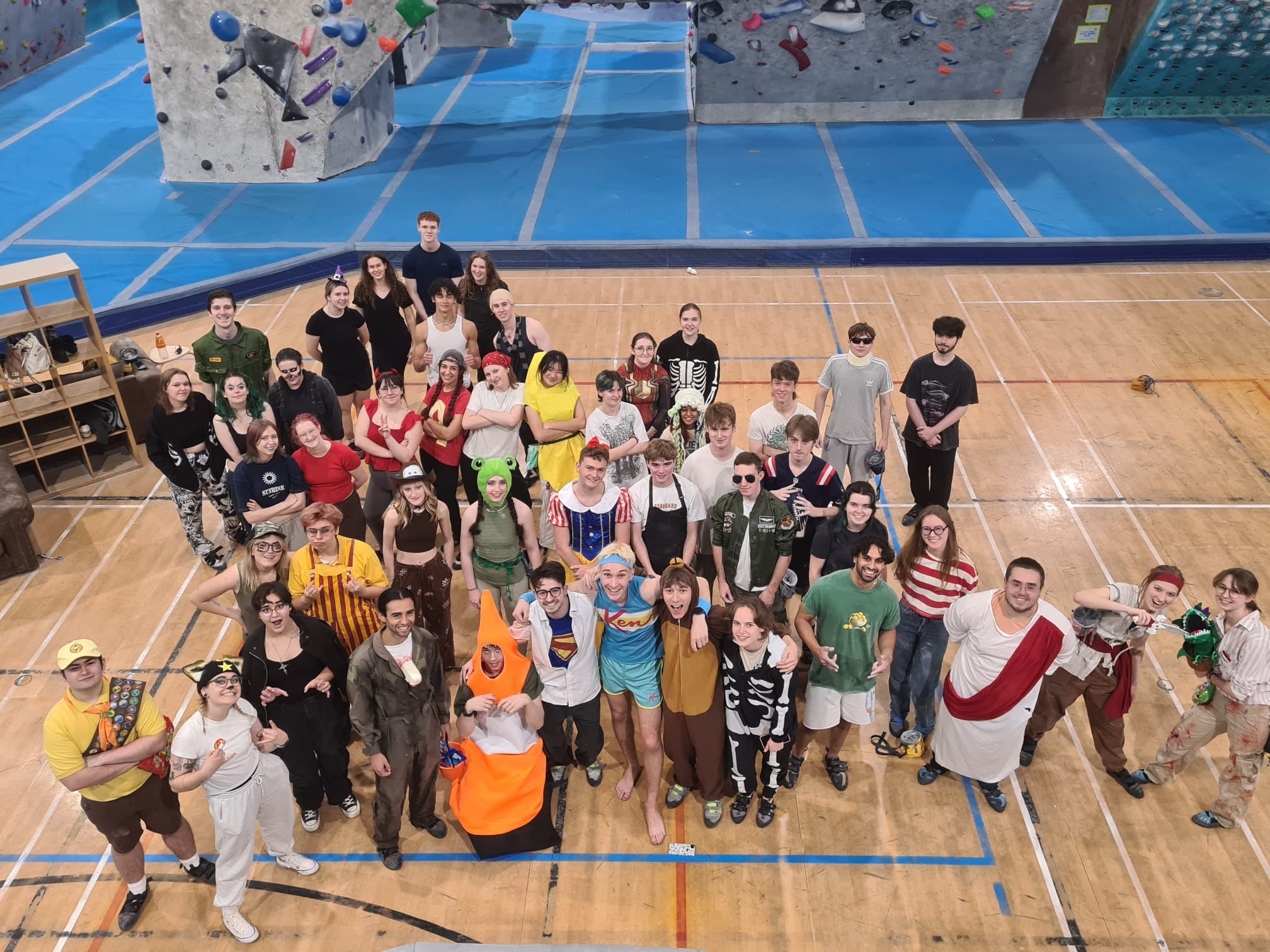 Large group of people in fancy dress costumes standing together on wooden gymnasium floor with blue court markings and climbing wall visible.