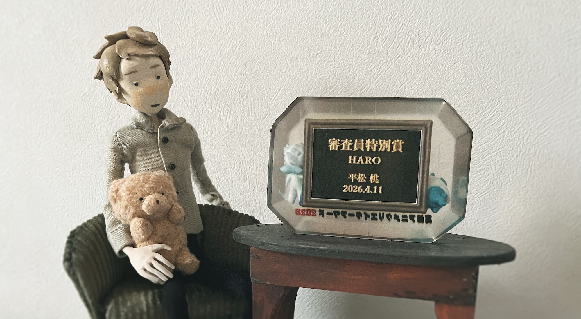 Stop motion animated model of a person sitting in a small armchair holding a teddy bear. They are sat next to a table with an award on it, written in Japanese script.