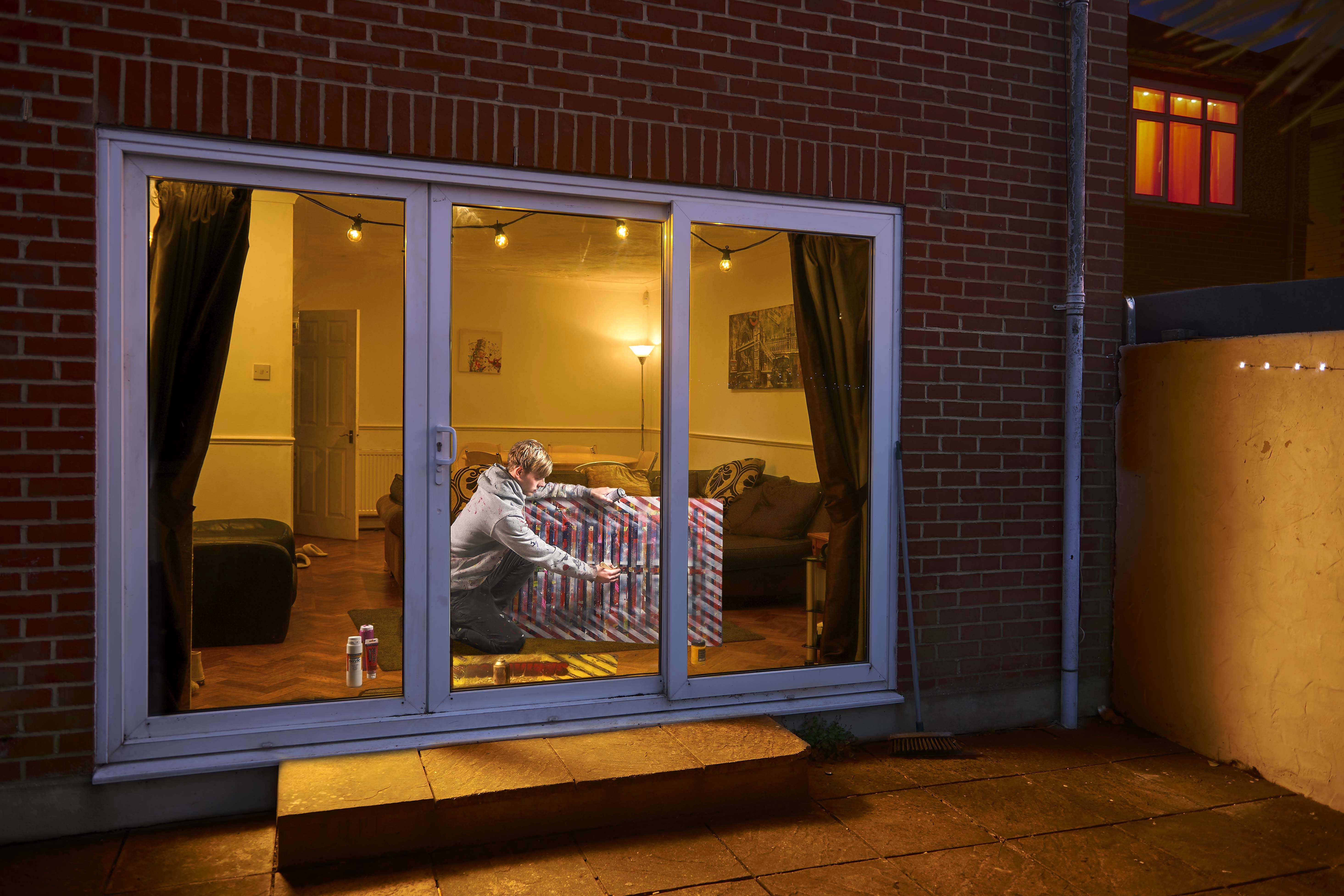 Student photographed in a living room from outside a house. The student is knelt down by a painting and working.