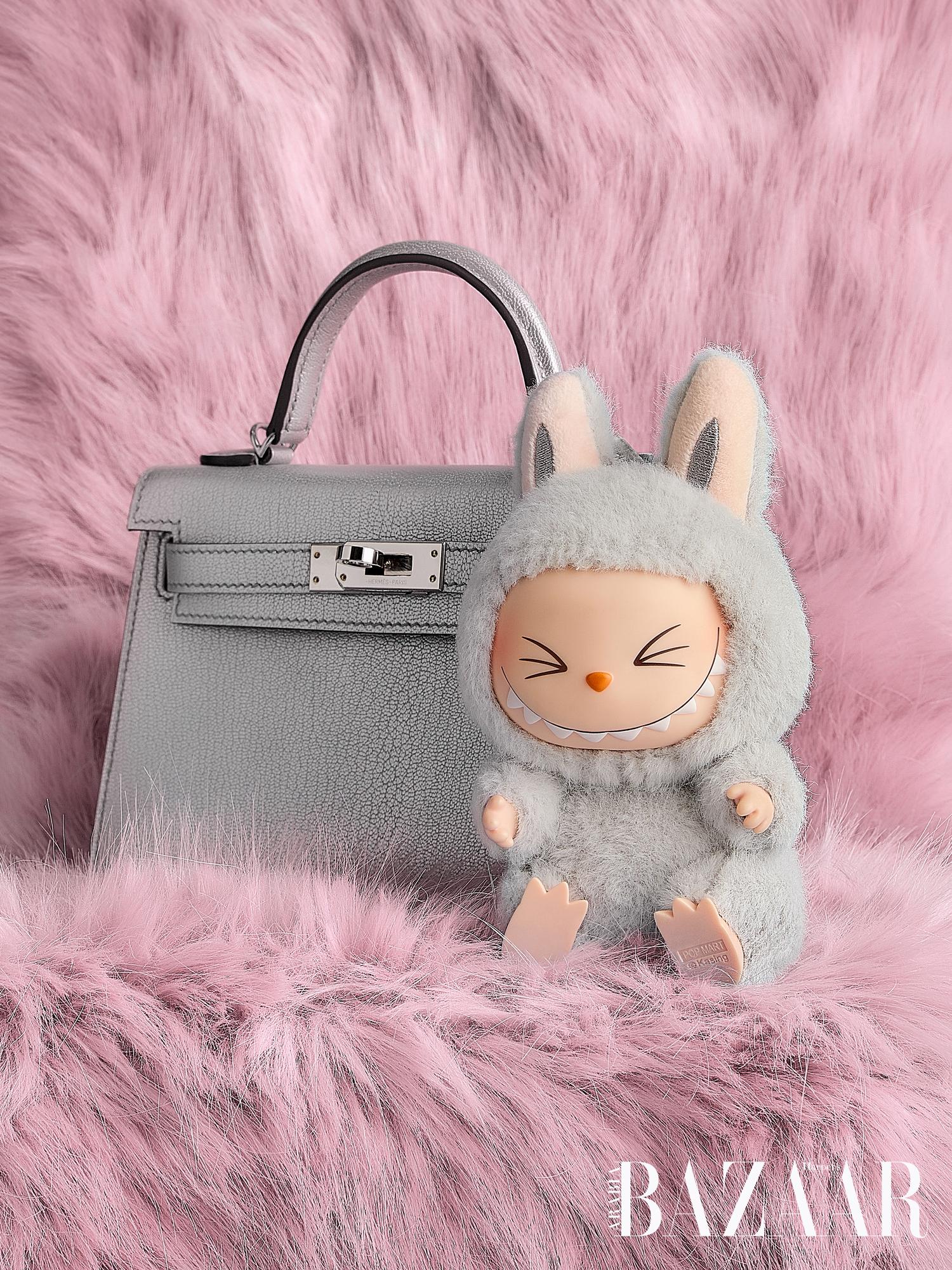 A grey leather handbag with a structured design is positioned next to a small toy figure dressed in a bunny costume, sitting on a pink fluffy surface. The background features a soft pink fur texture, enhancing the playful aesthetic.