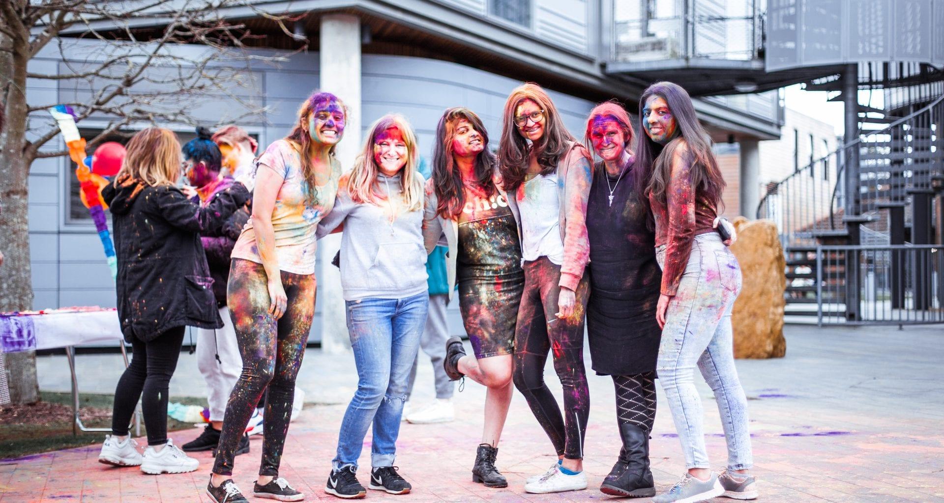 Group of people covered in coloured paints line up in a courtyard and pose for a photo.