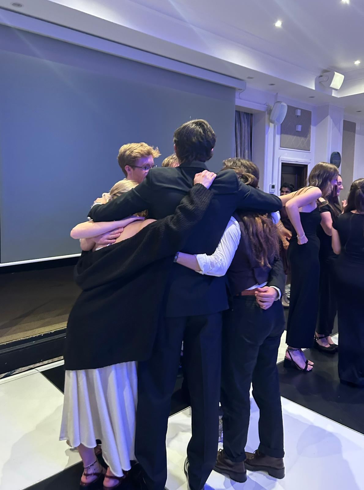 Group of people in dark clothing embracing in a circle huddle inside modern room with purple lighting and white ceiling.