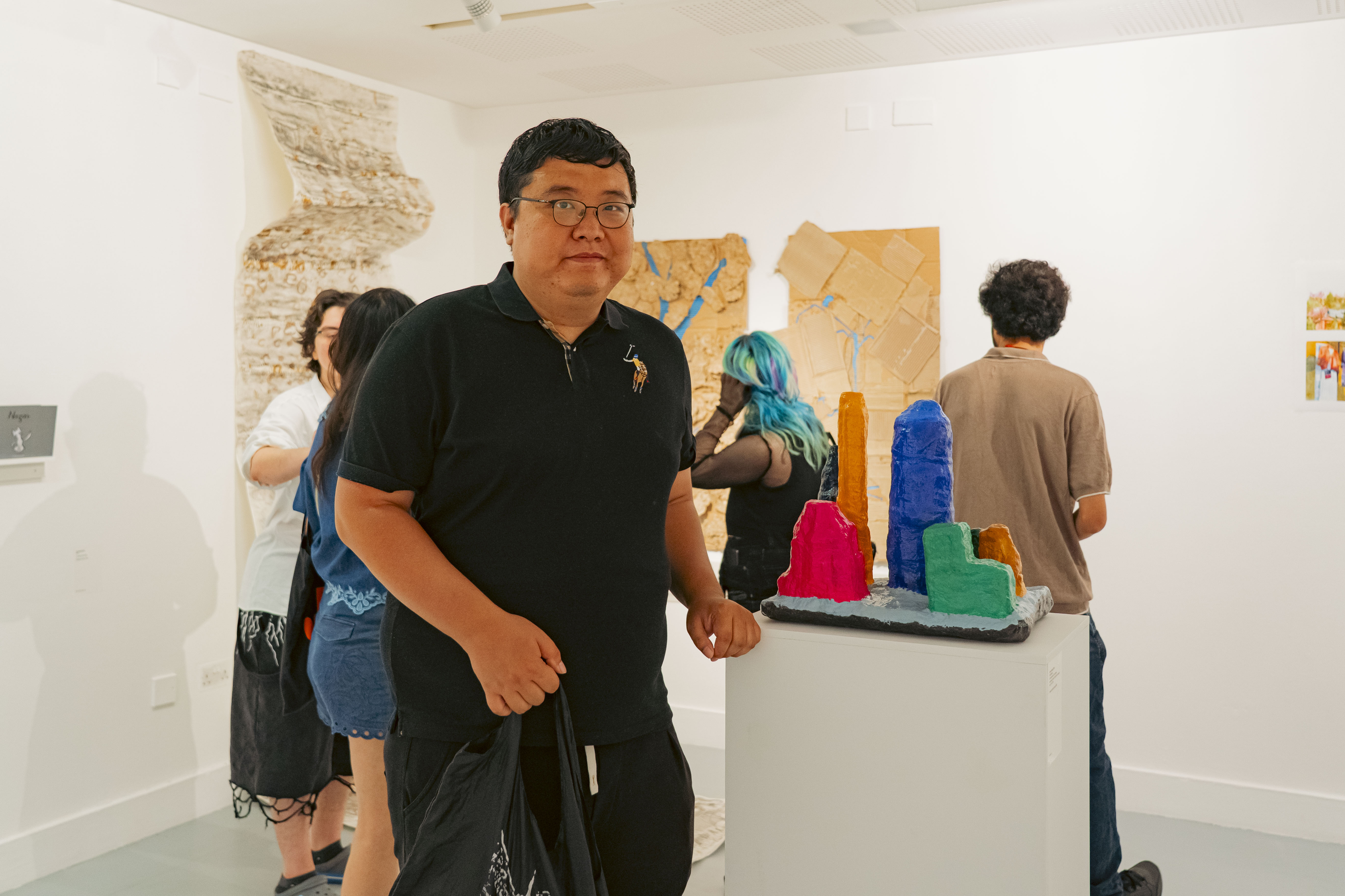 A man in a black polo shirt stands in an art gallery, with several visitors and colourful sculptural pieces on white plinths in the background.