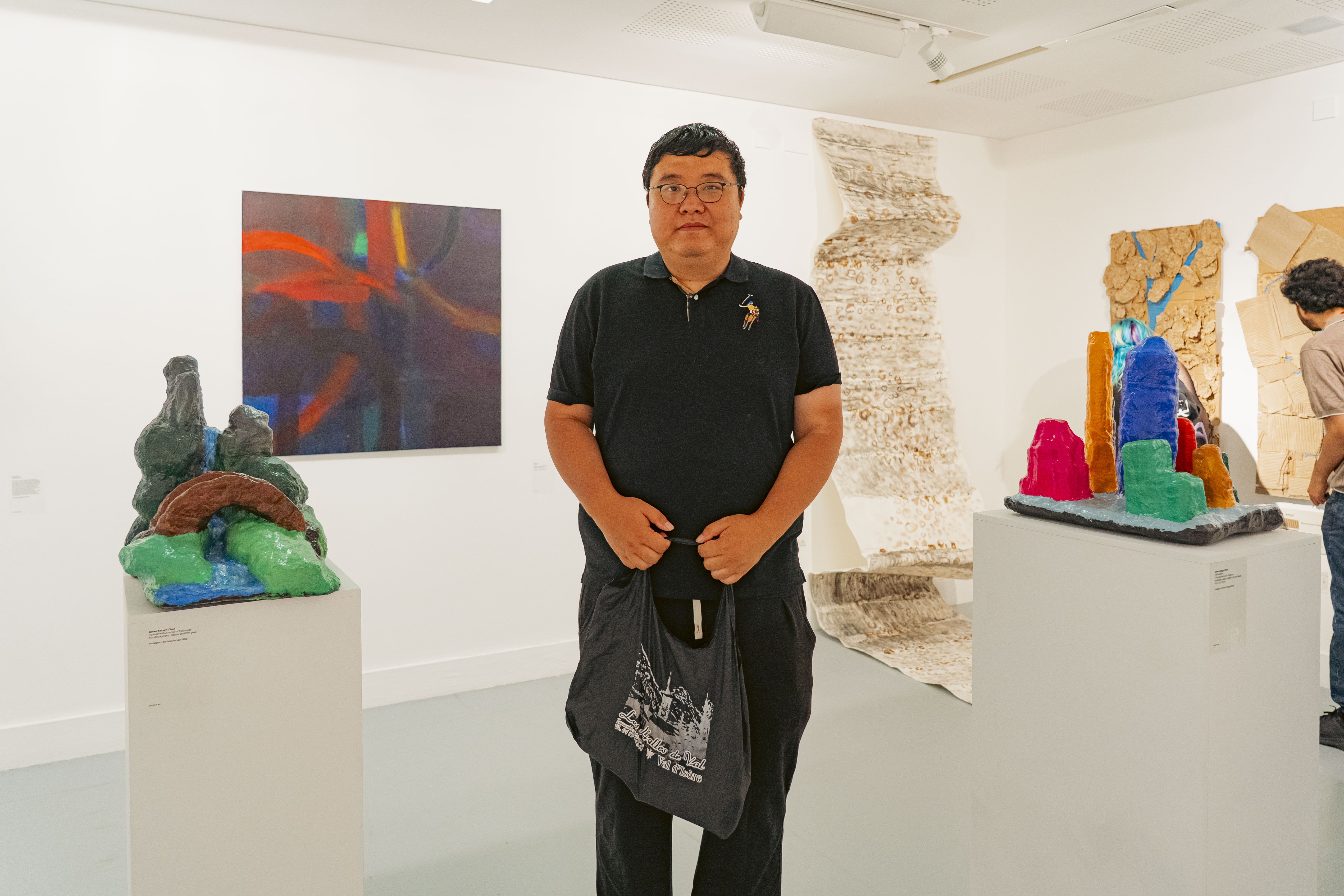 A man in a black polo shirt stands in a white gallery, with colourful sculptures on pedestals and abstract artworks on the walls.