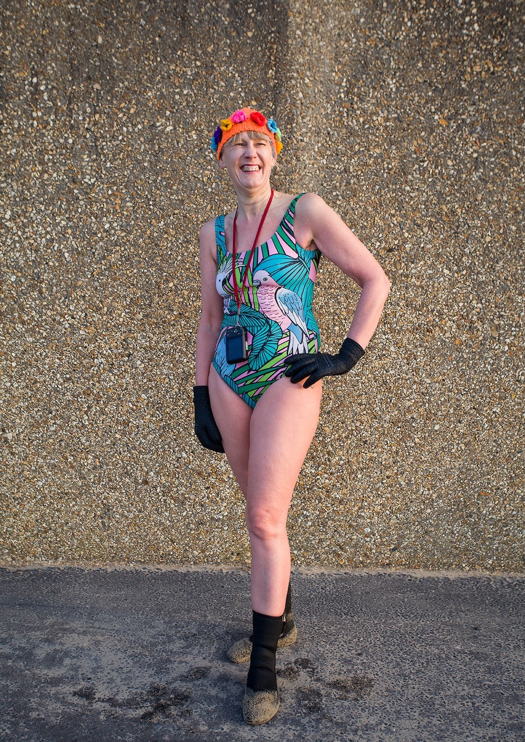 Woman in colourful tropical print swimsuit and orange swimming cap standing on beach, wearing black gloves and boots, smiling.