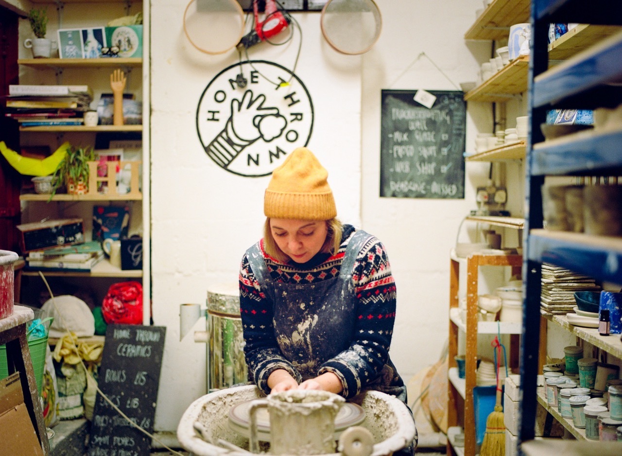 Person in yellow beanie and patterned jumper working at pottery wheel in cluttered studio with shelves of supplies and circular logo on wall.