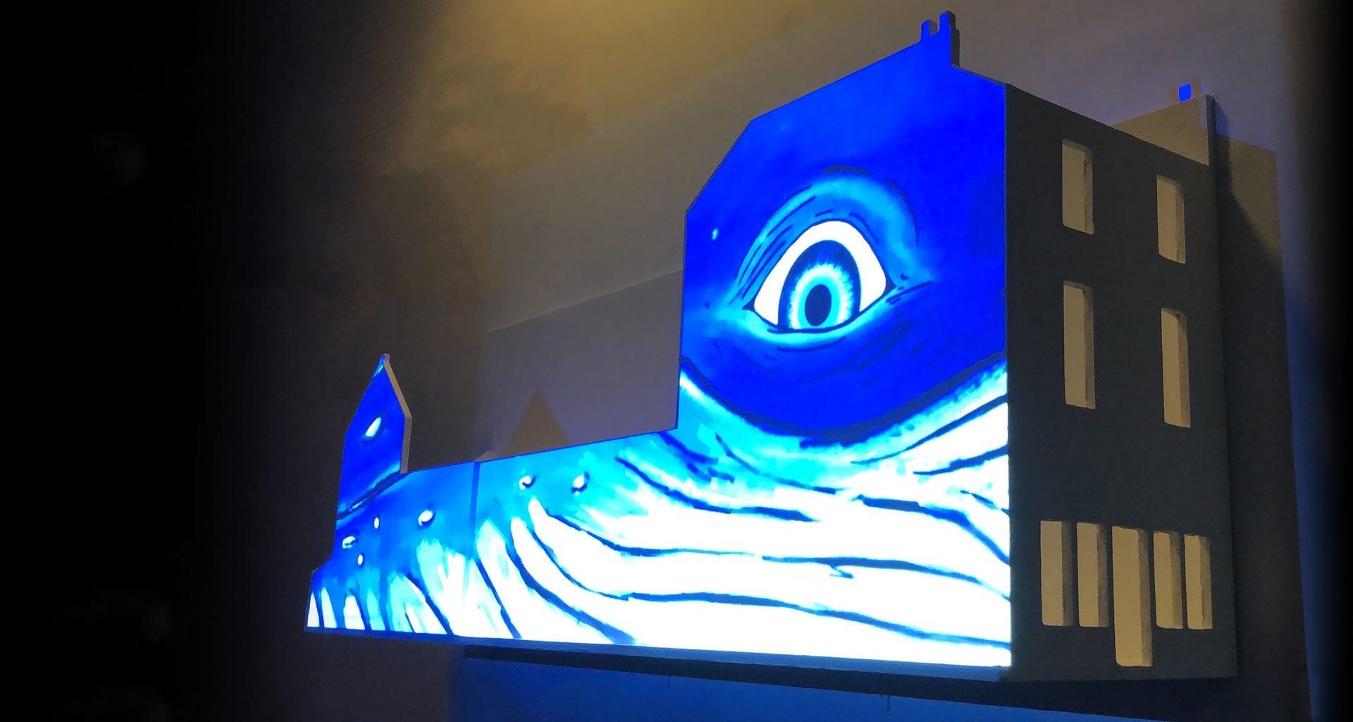 The image of a monster's face, including a large eye, is projected onto the side of a model building.