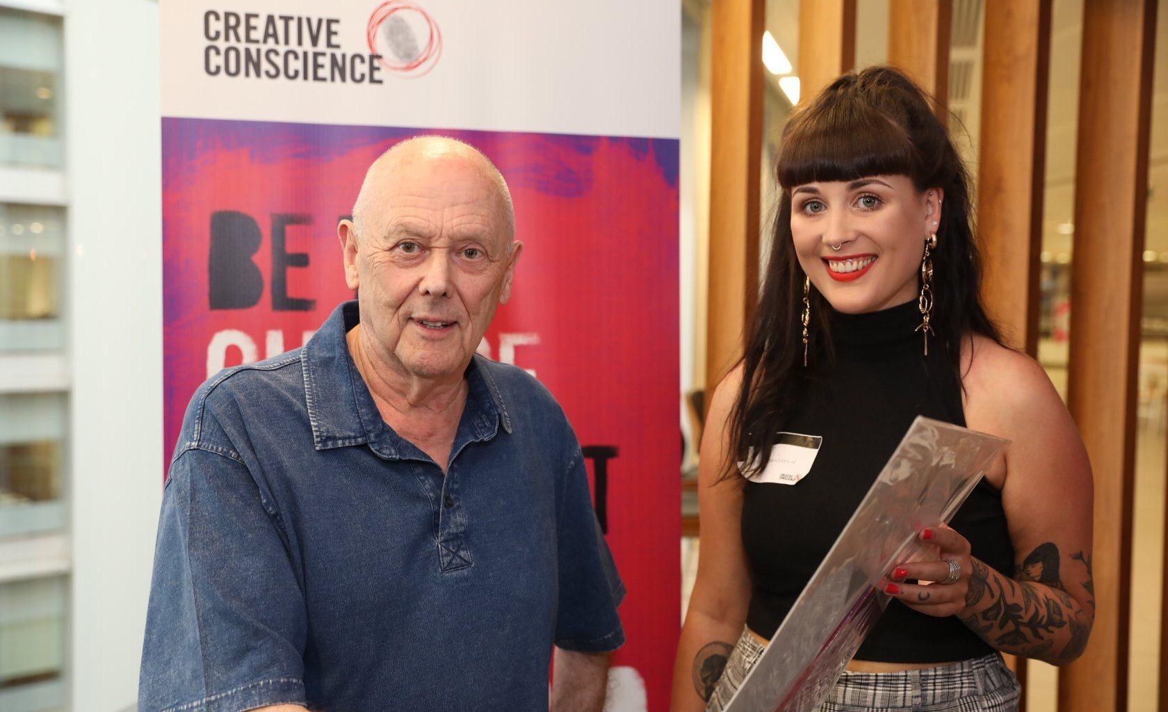 Two people stand and pose for a photo in front of a banner. The words 'Creative Conscience' appear at the top of the banner.