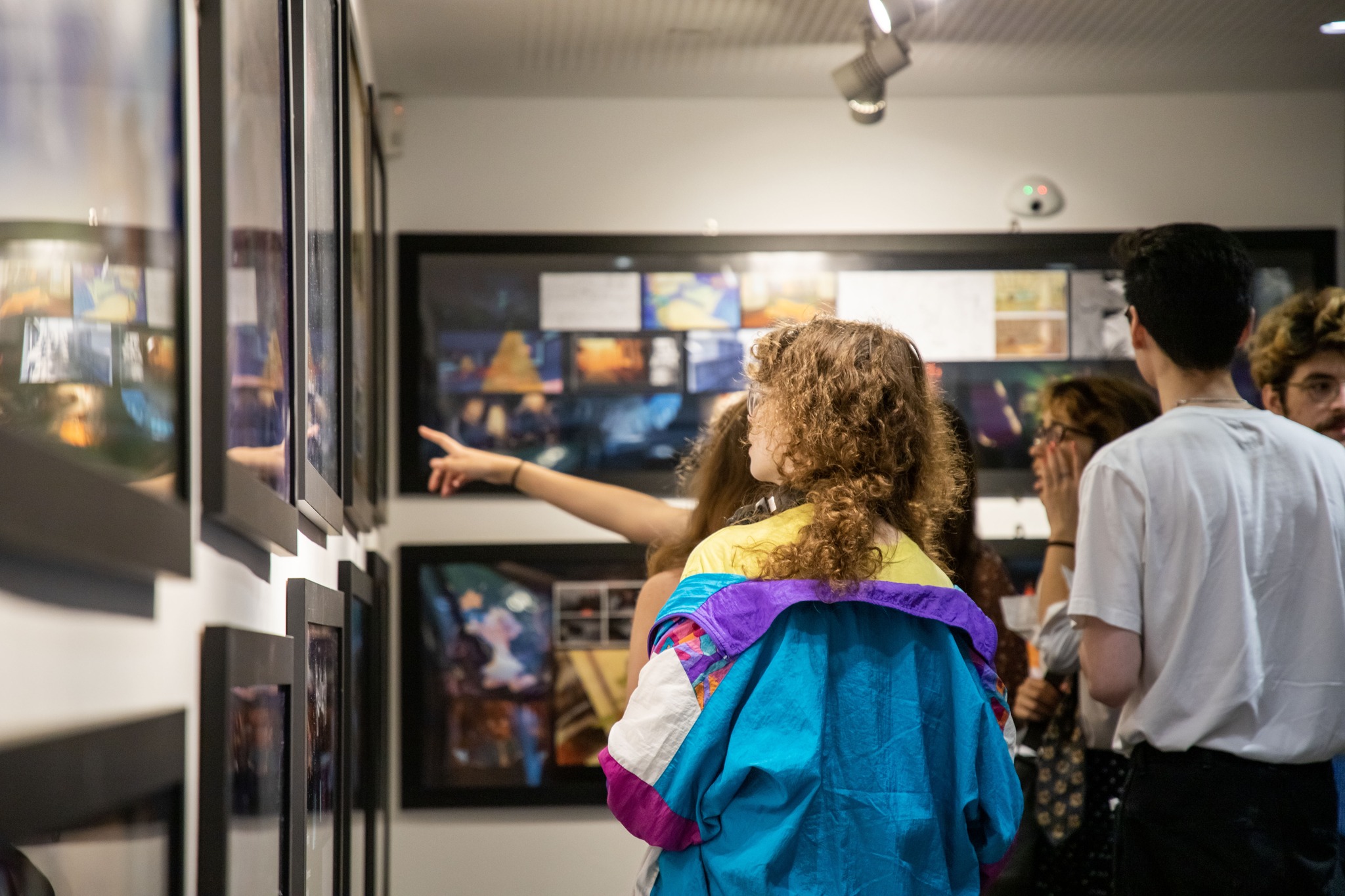 Child with blue coat and frizzy hair looking up at black framed artworks, person behind them pointing upwards.