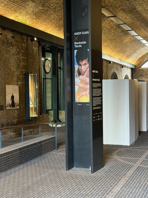 Black pillar featuring portrait image of Prince in yellow, taken by Andy Earl, illuminated brick corridor in the background.