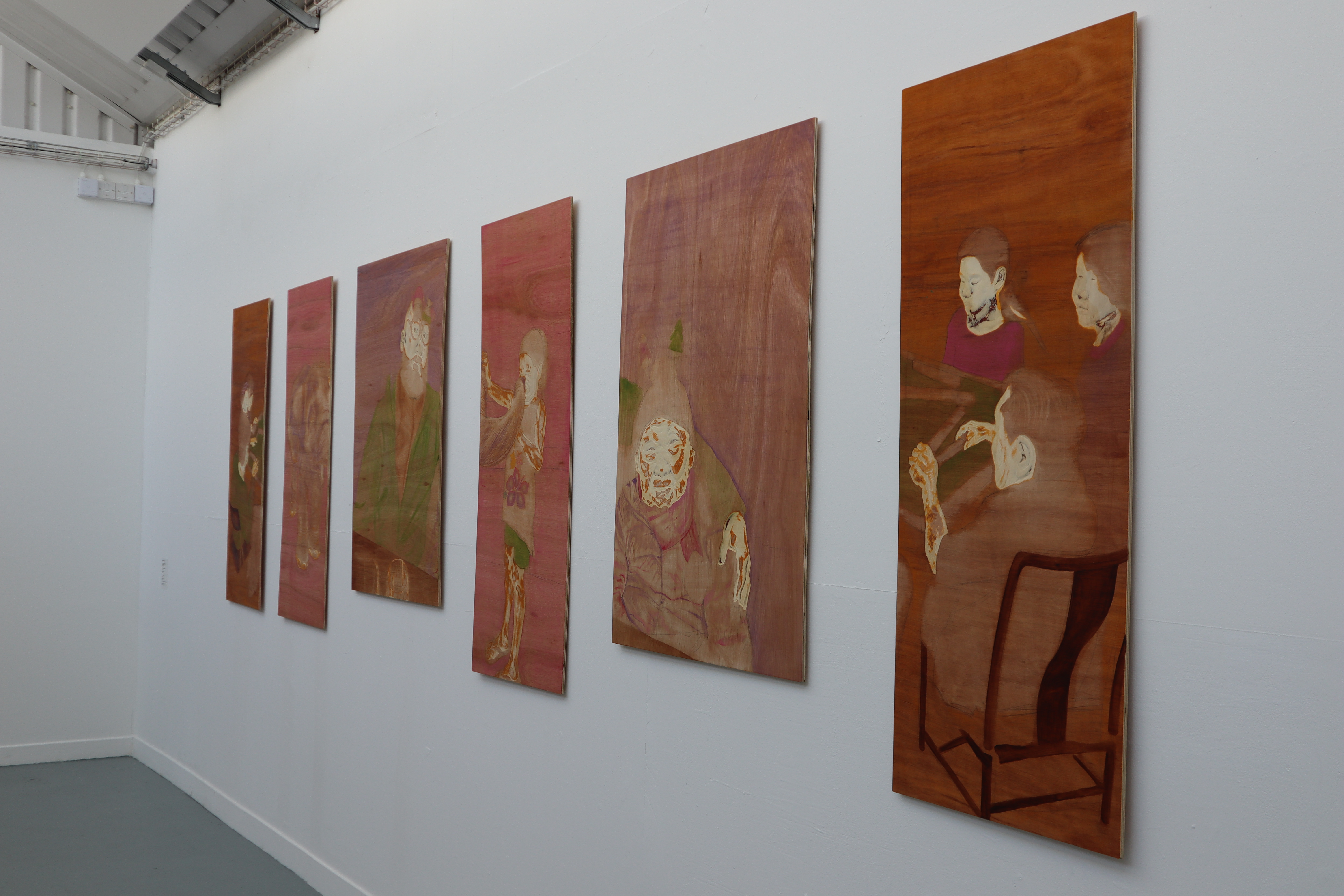 Six tall wooden art panels hang in a row on a white gallery wall, with painted figures and warm brown tones, in a spare room with a grey floor.