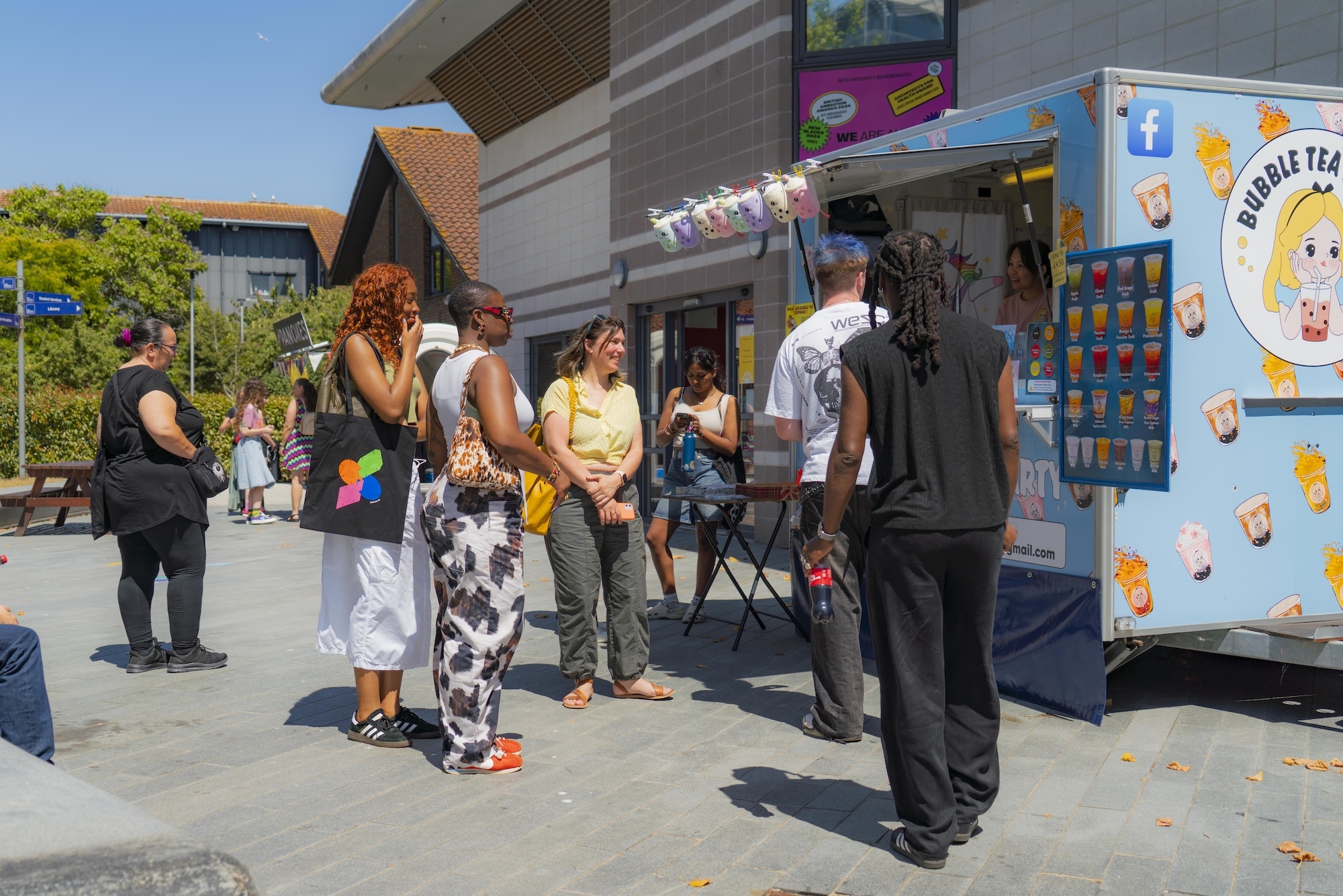 People waiting at a light blue bubble tea stand, one with colourful black AUB tote bag, sunny day.