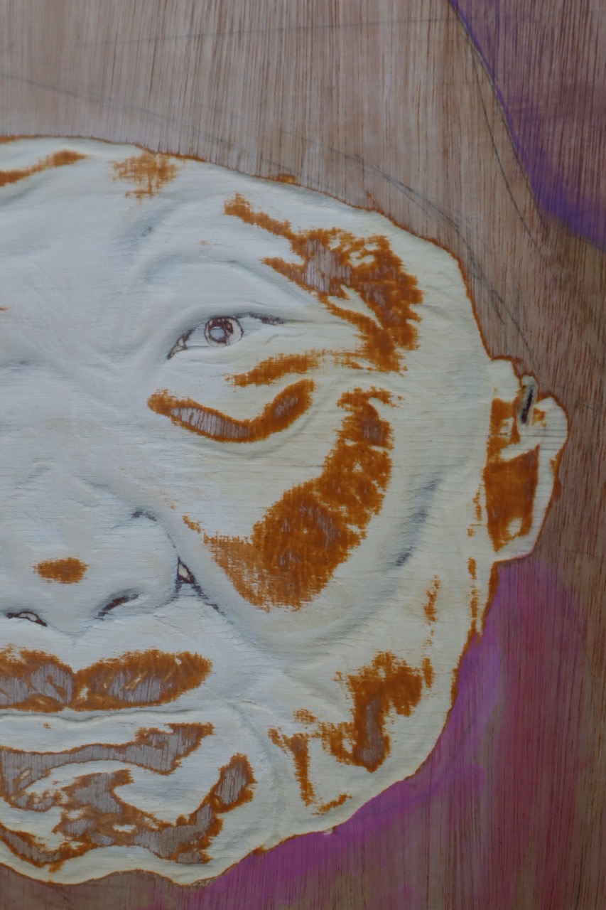 An abstract close-up of a painted human face in white and brown tones on a wooden surface, with purple shading in the background.