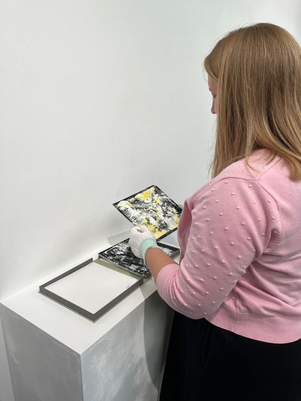 A woman in a pink cardigan handles a small open box and artwork on a white pedestal against a plain wall.