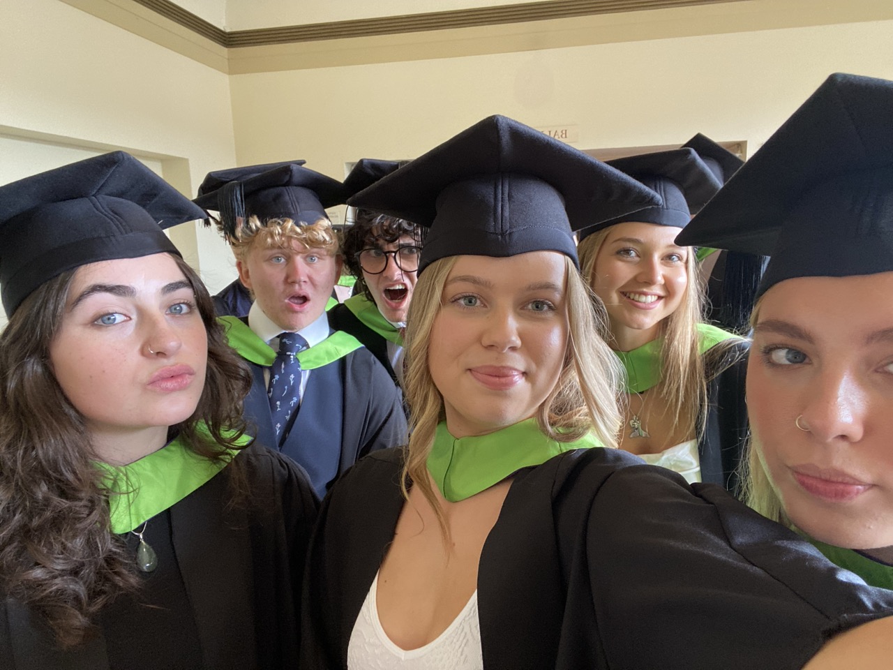 Six graduates in black caps and gowns with green collars posing for a group selfie indoors, with beige walls in the background.