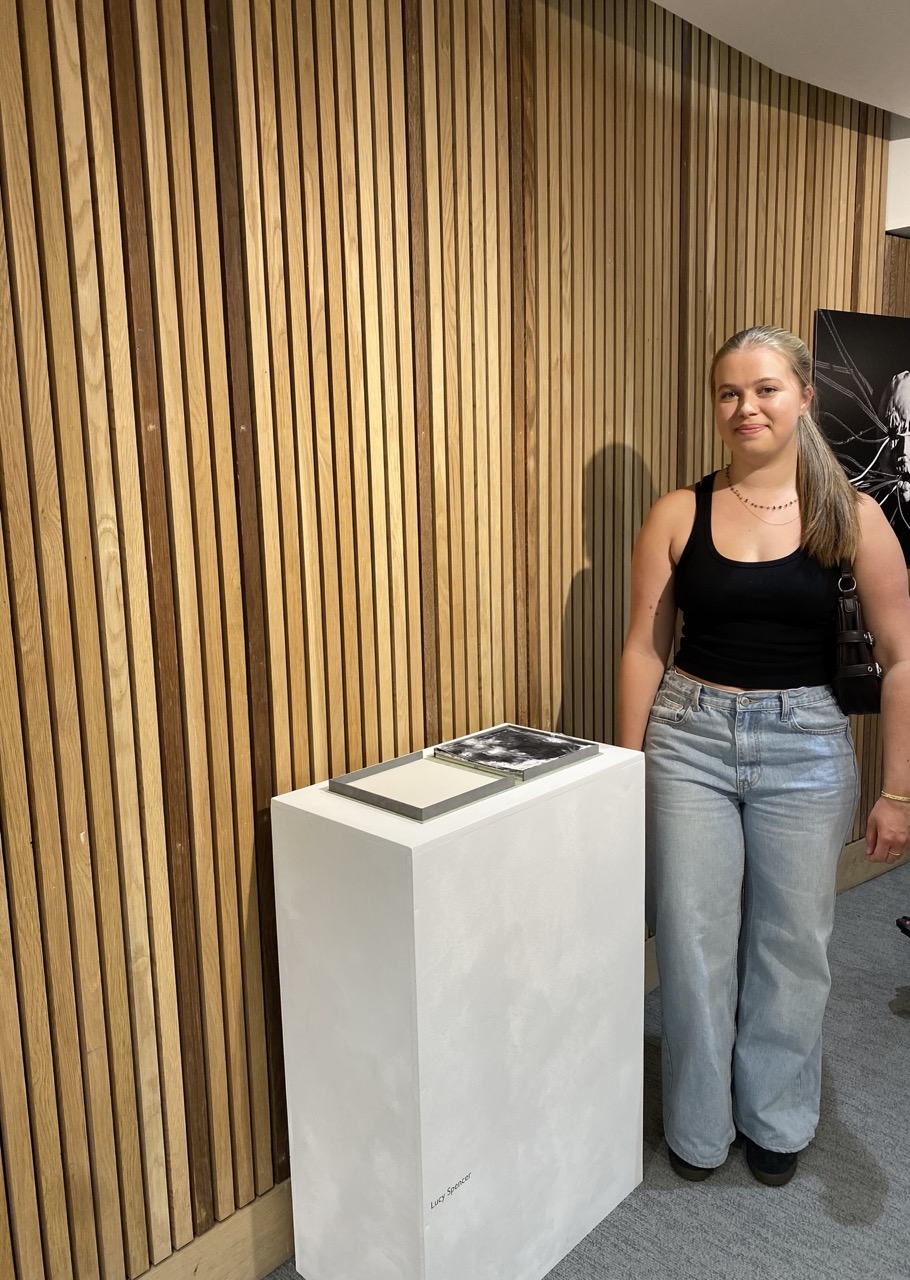 A woman in a black sleeveless top and light blue jeans stands beside a white display pedestal in front of a vertical wooden slat wall.
