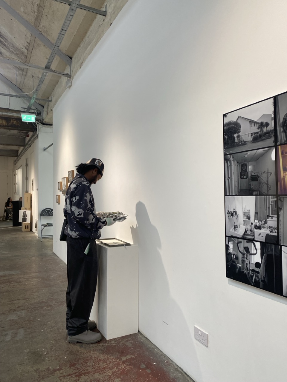 A man in a dark patterned jacket and cap examines items on a small white pedestal beside a gallery wall, with black-and-white photo panels hanging nearby.