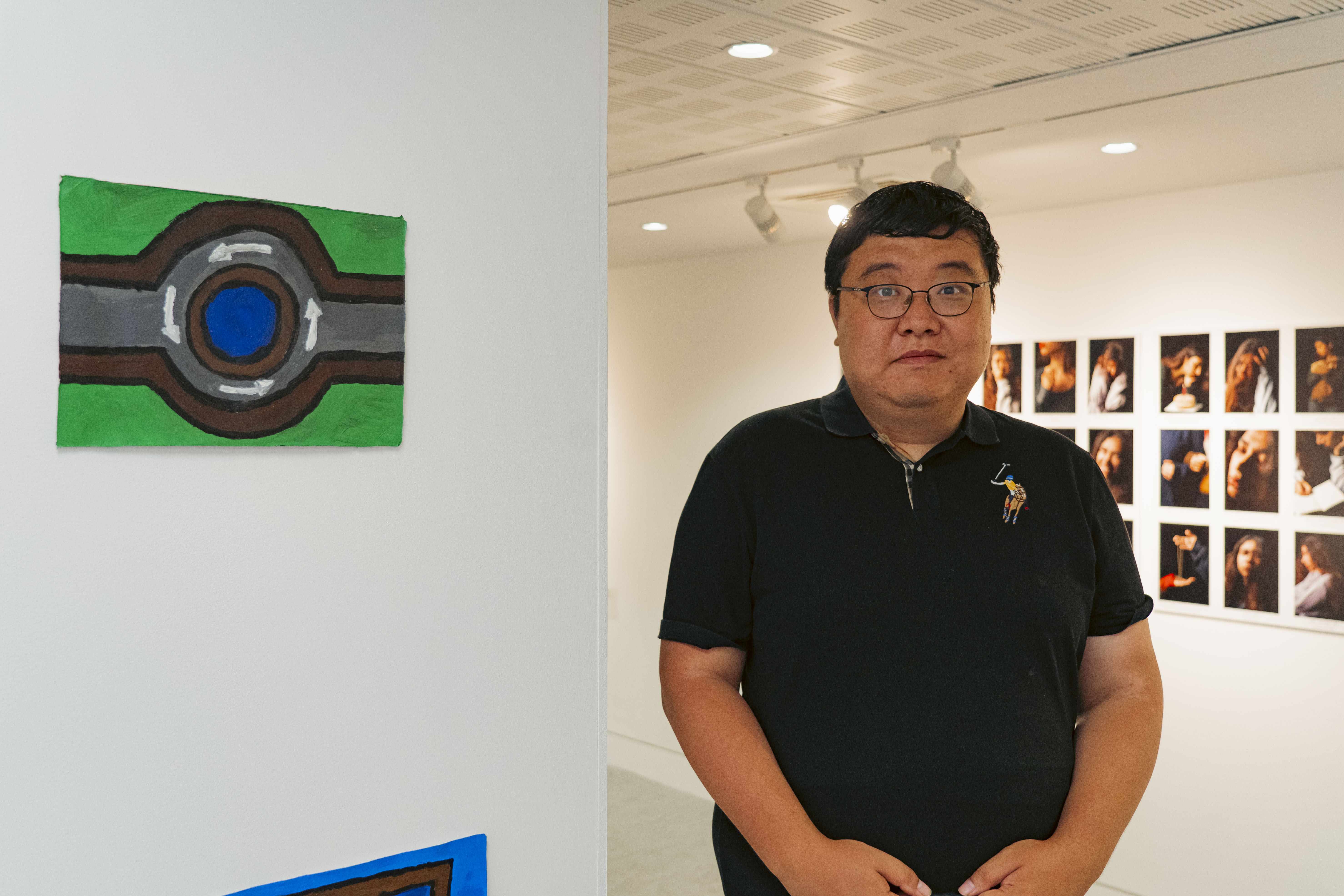A man in a black polo shirt stands in an art gallery beside a colourful abstract painting on a white wall, with framed photographs displayed behind him.