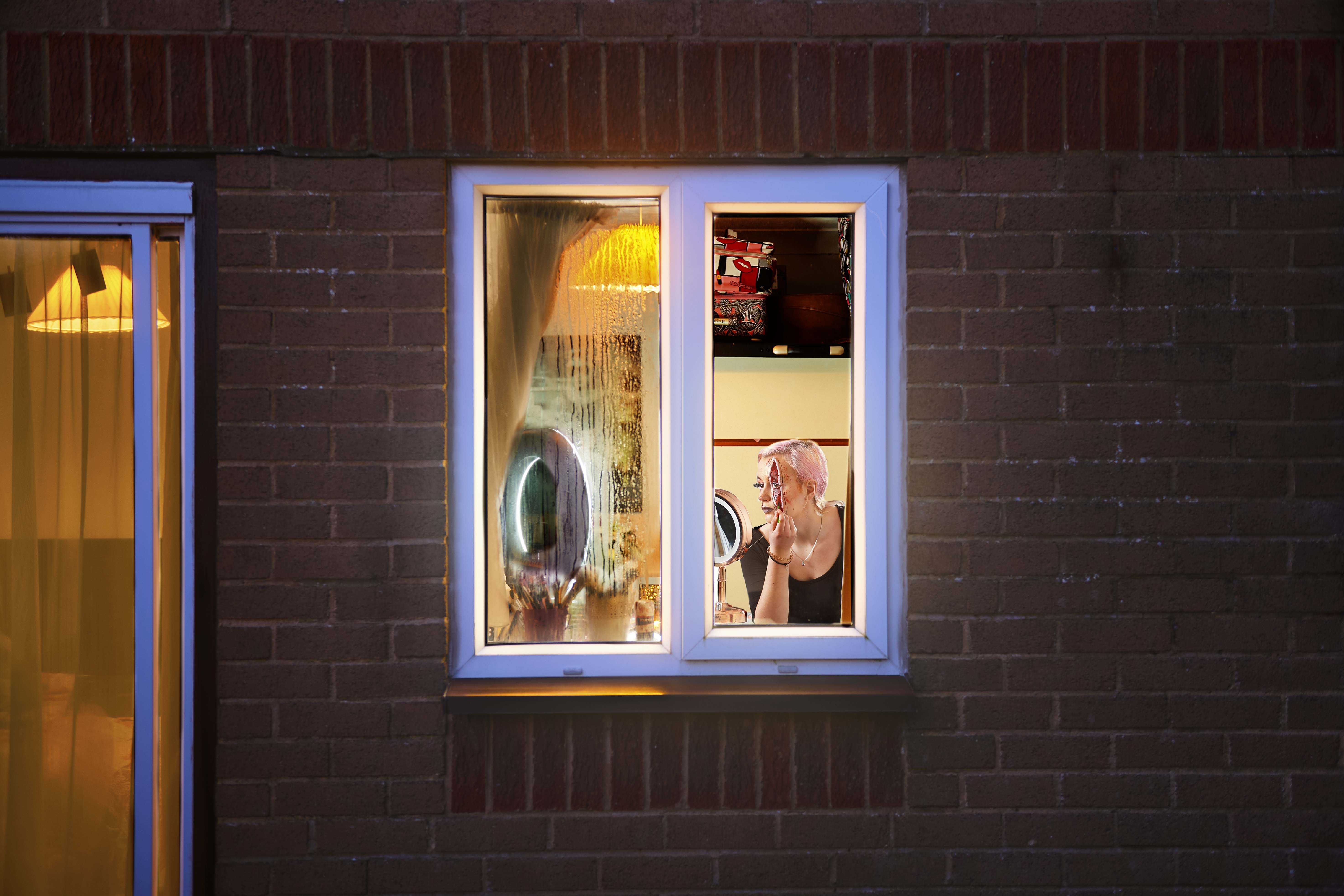 A student viewed through a small window from outside a house. The student is applying a prosthesis while looking in a mirror.