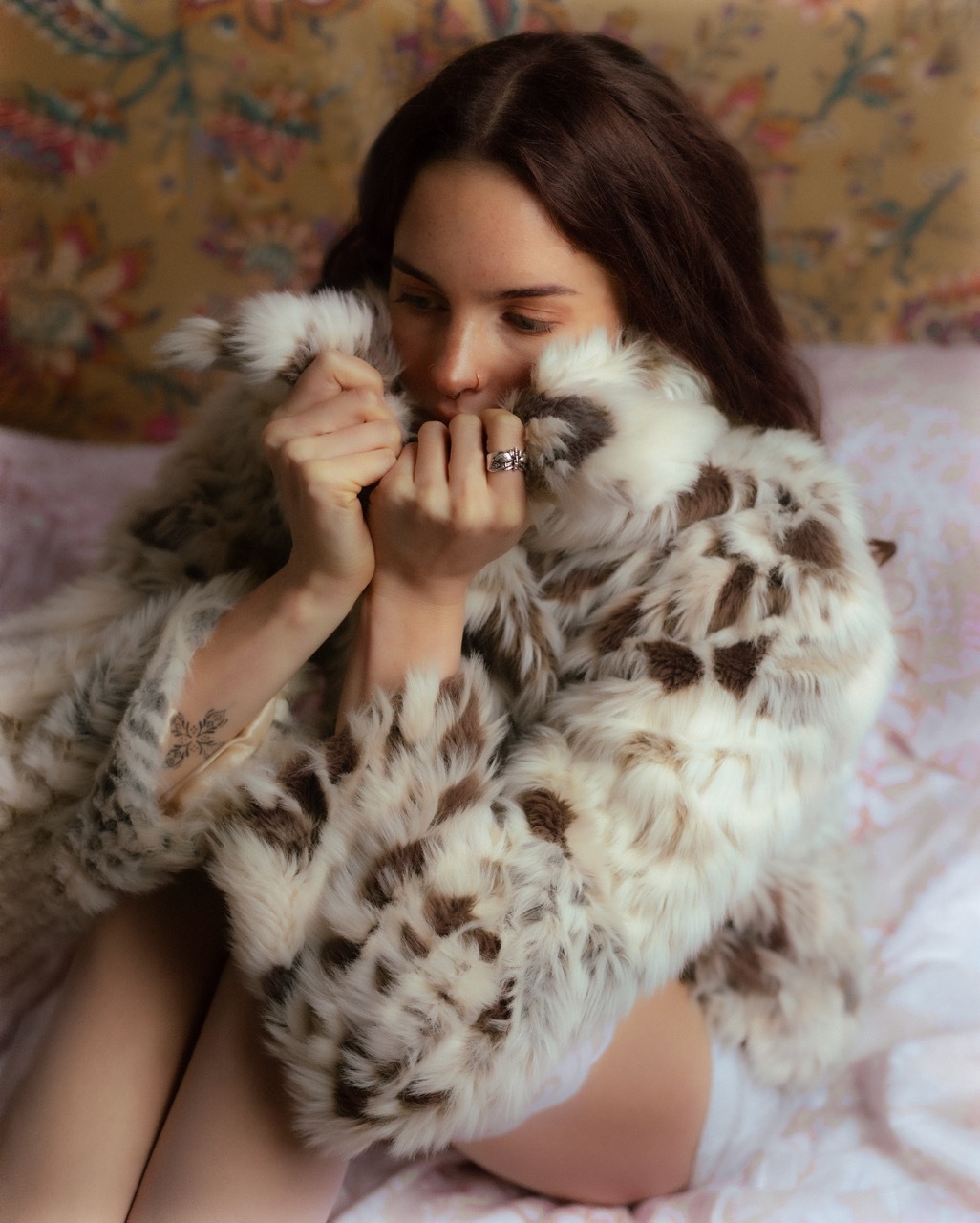 A model wearing a faux-fur coat on a pink bed and floral background.