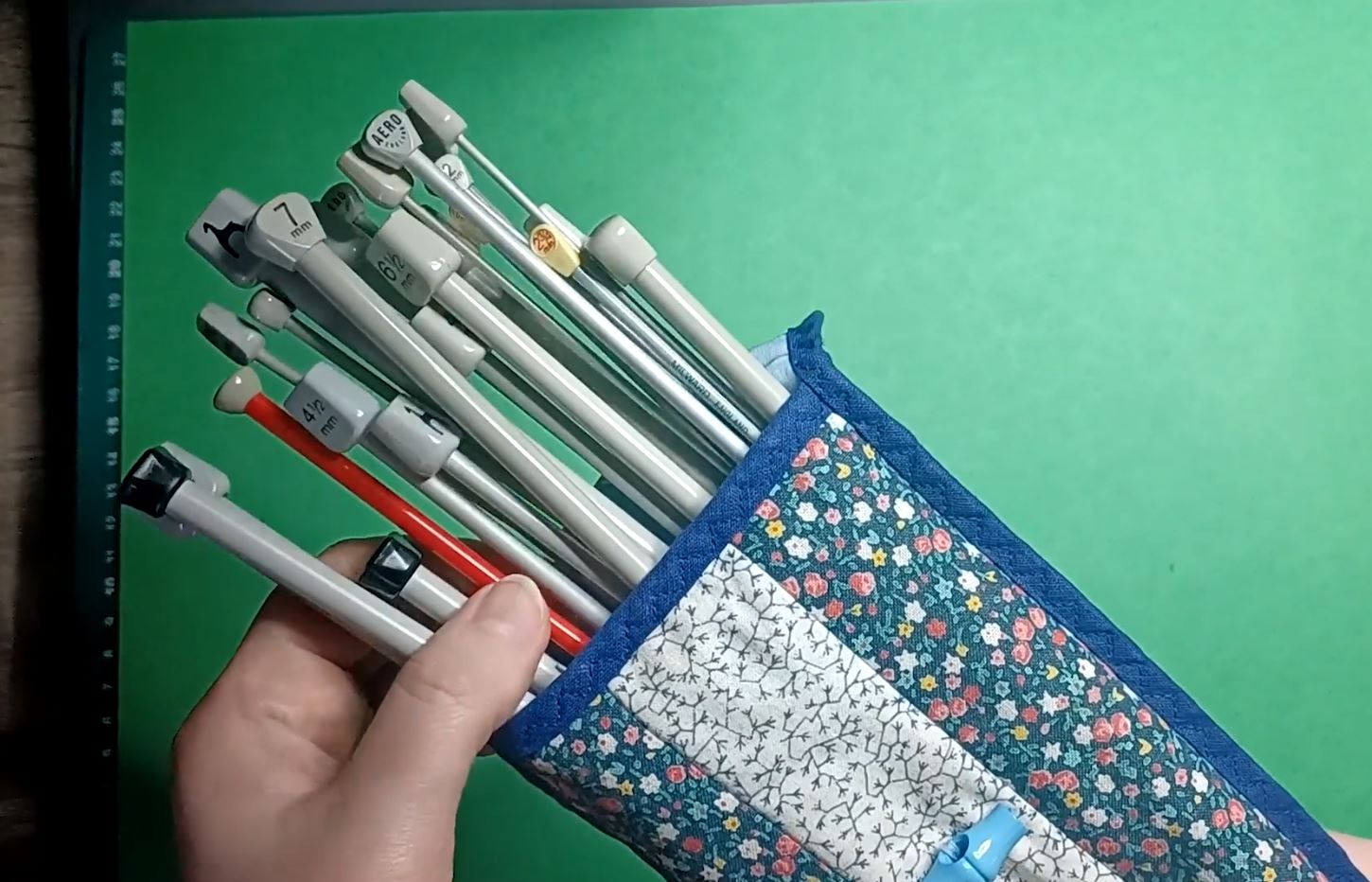 A hand holds a floral fabric pencil case filled with mechanical pencils, rulers and pens against a green background.
