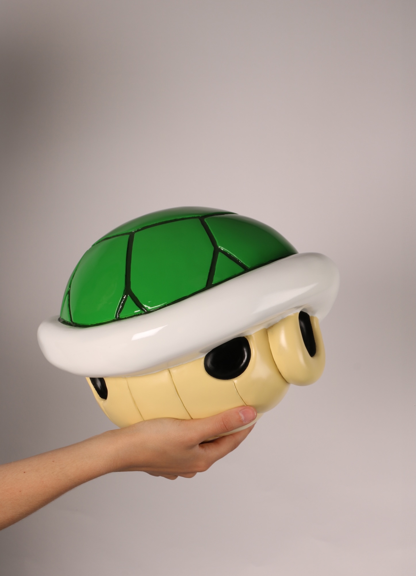Hand holding green Koopa Troopa shell model with yellow base, white rim, and black holes against grey background.