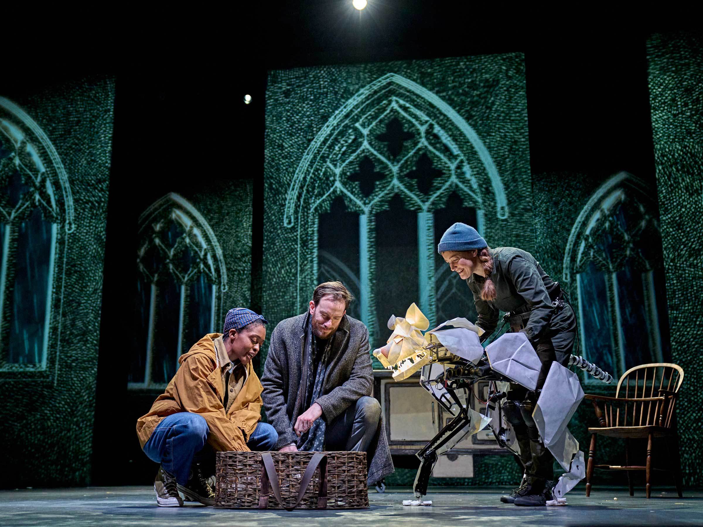 Two actors in costume crouching down by a basket on a stage. A third person operates a hyena puppet.