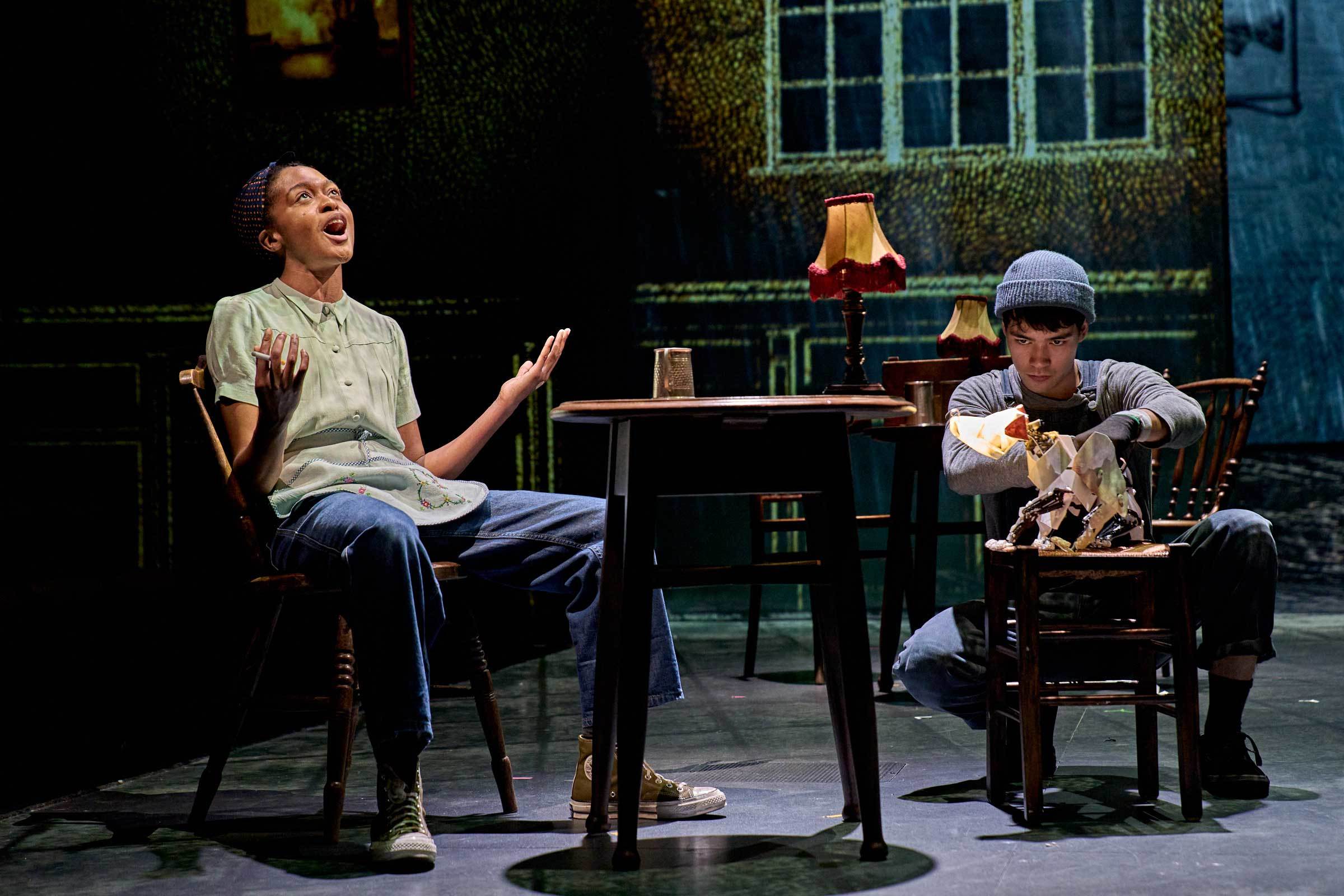 Two actors in costume sit opposite sides of an old-fashioned table on a stage.