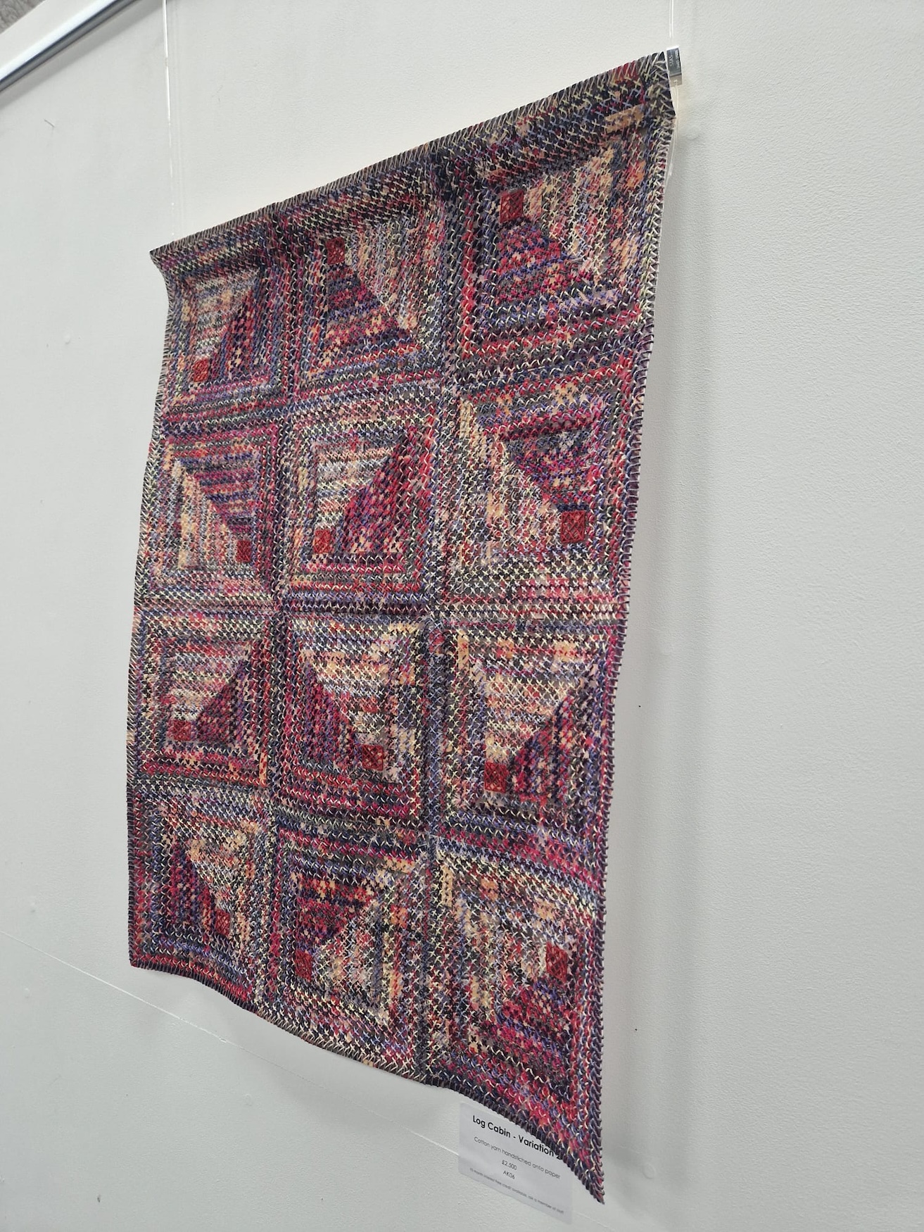 A textile piece displayed on a wall, featuring a patchwork design with twelve squares arranged in a grid. The squares are composed of various fabrics in shades of red, purple, and cream, creating a textured and colourful pattern.