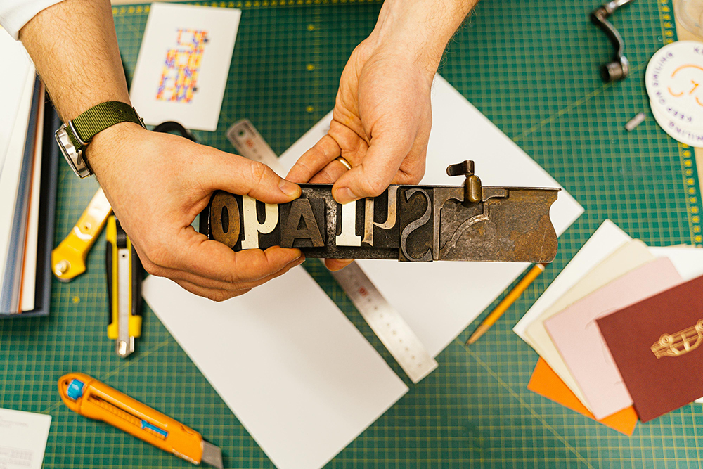 Hands holding metal letter stamps above white paper on green cutting mat, surrounded by craft supplies and papers.