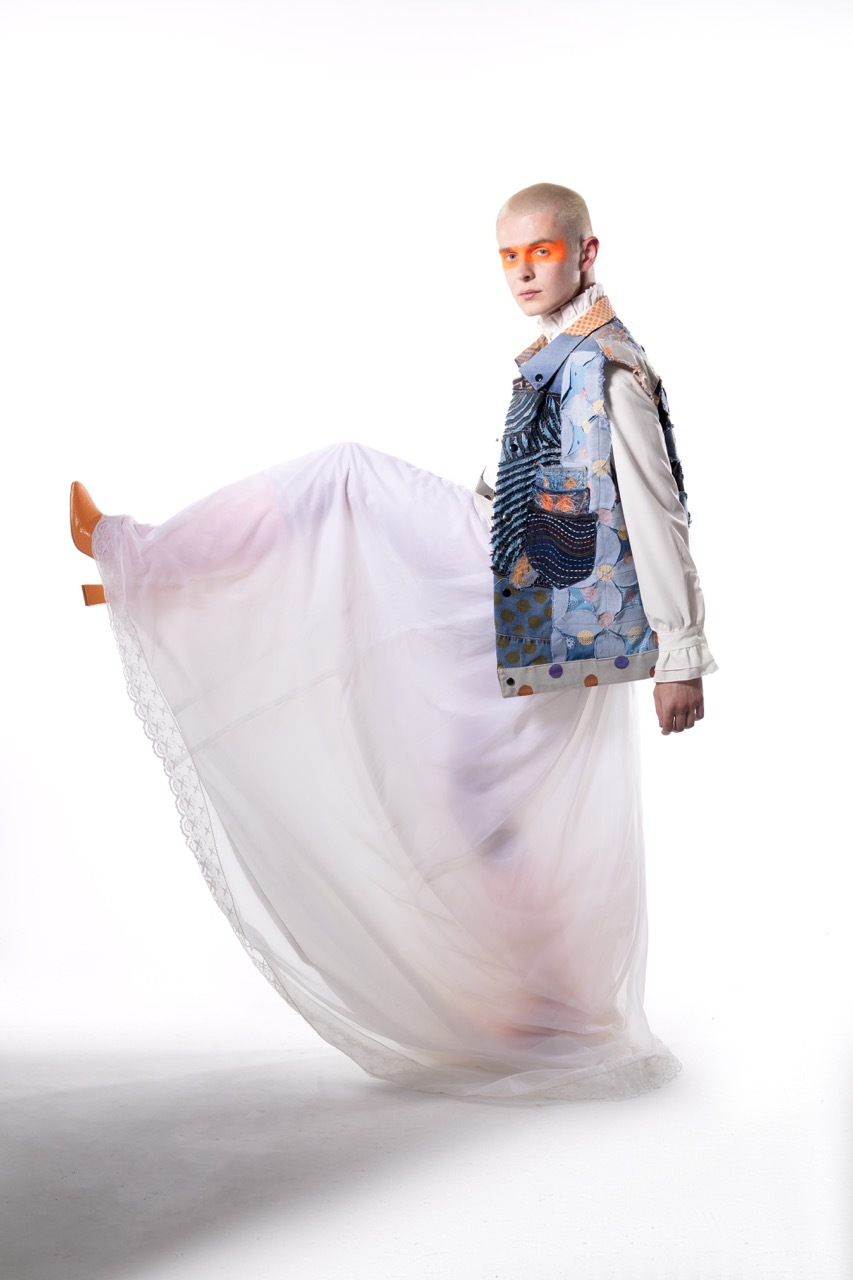 Person with orange face paint wearing patterned blue vest over white shirt, flowing white fabric around legs, white background.