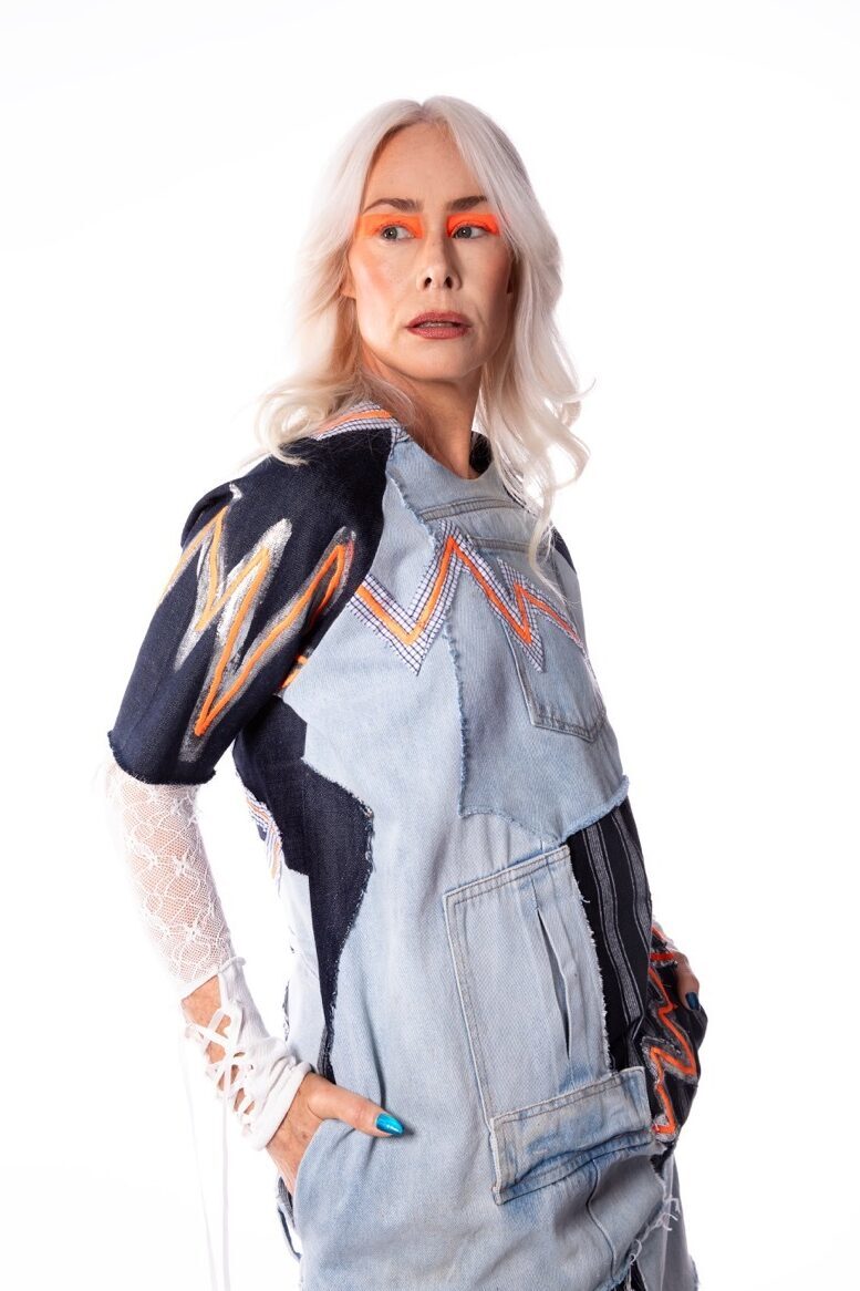 Person with platinum blonde hair wearing light blue denim jumpsuit with colourful geometric patterns on sleeves, orange face paint.