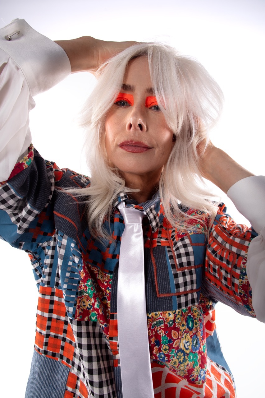 Woman with platinum blonde hair and red eyeshadow wearing patchwork jacket with gingham, floral, and geometric patterns and a silver tie.