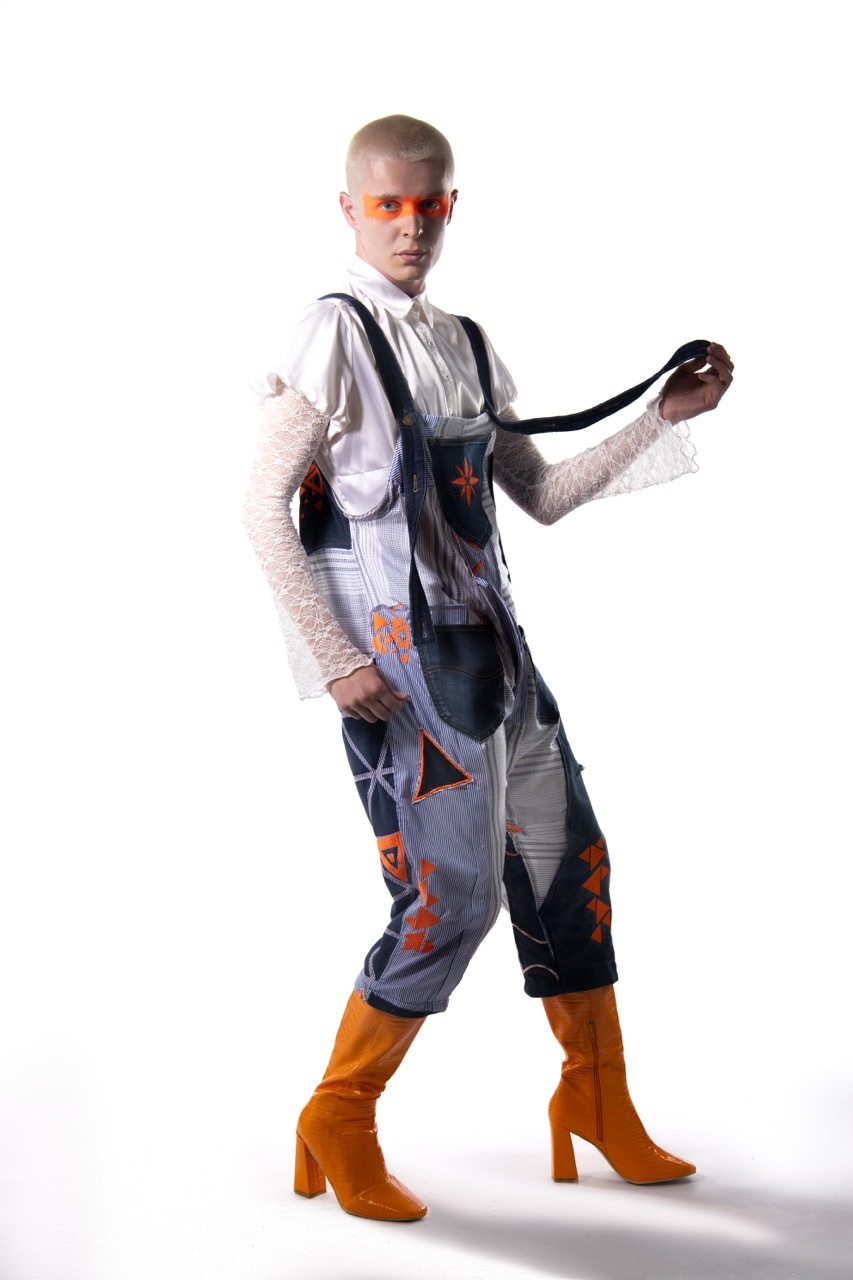 Person in white top and grey patterned dungarees with orange boots, orange sunglasses, posing with one arm extended on white background.