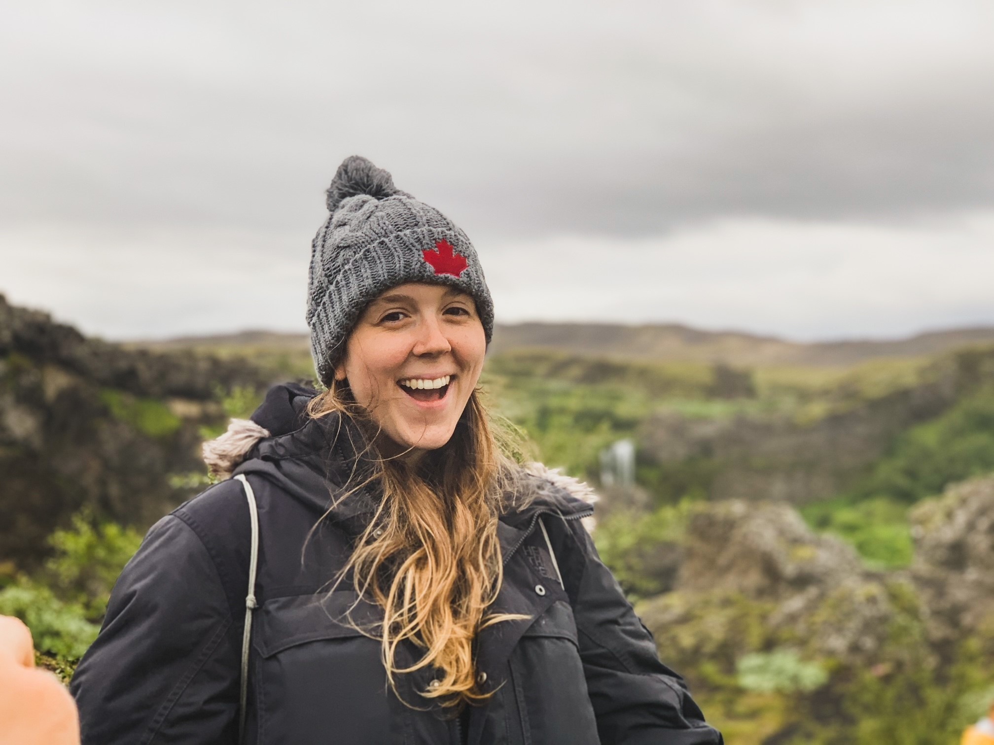 Woman in grey knit hat with red logo and black jacket laughing outdoors, with green hilly landscape and cloudy sky behind her.