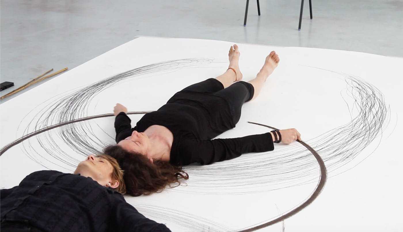 Two women lying down on top of a piece of paper with two incomplete circles drawn on it.