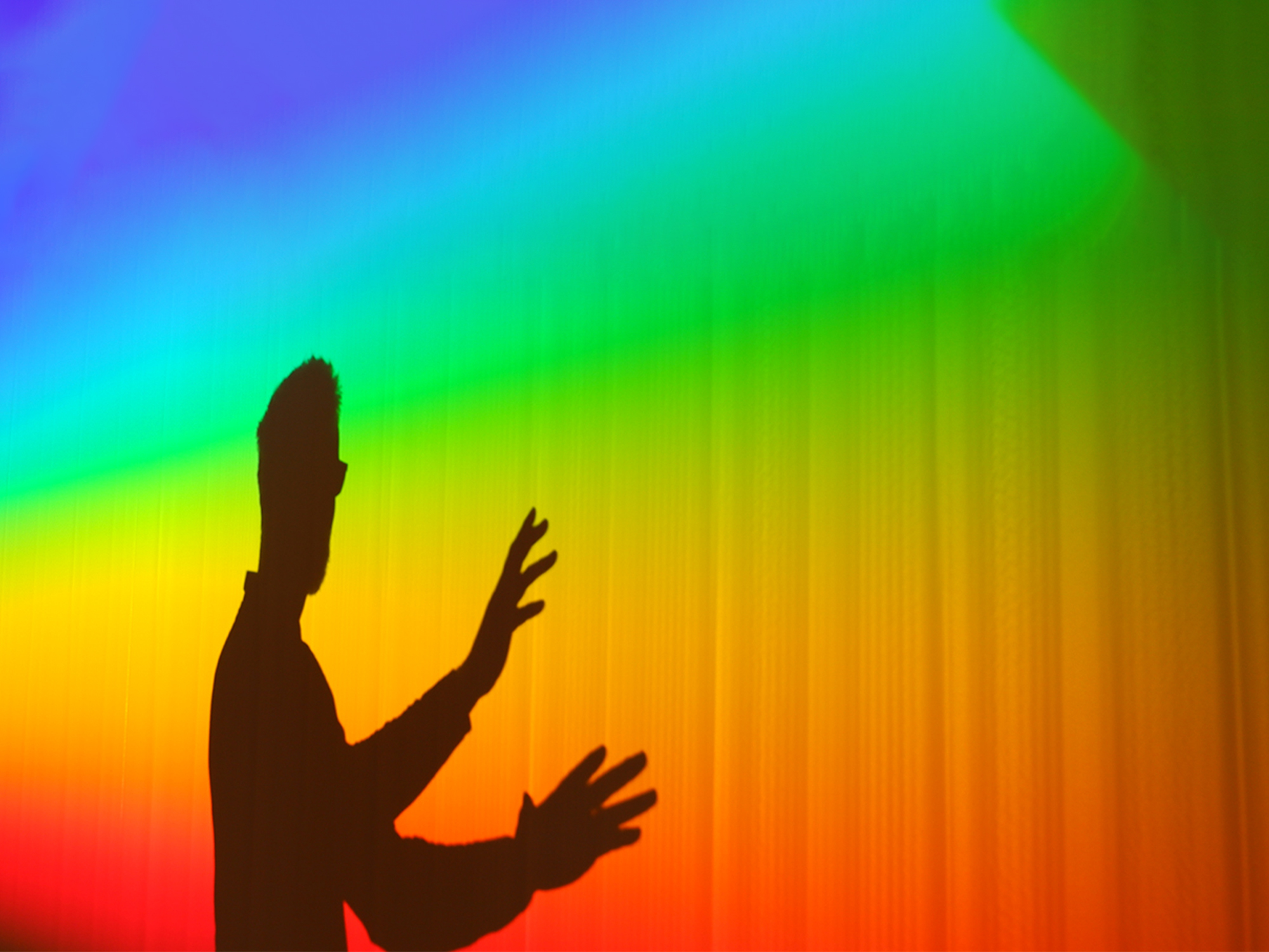 Black silhouette of person with raised arms against vibrant rainbow gradient background transitioning from purple at top to red at bottom.