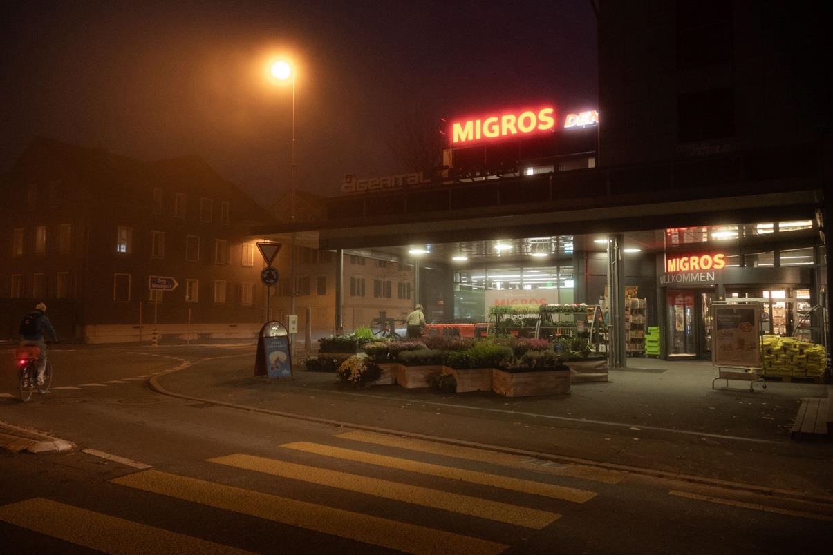 A Migros supermarket illuminated at night, with produce displayed outside. A cyclist is passing by in the foreground, while an urban environment with dim lighting and fog is visible in the background.
