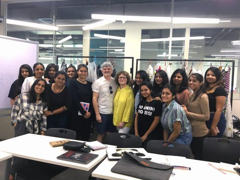 A group of Make-up for Media and Performance students gather in a conference room and pose for a photo with a pair of clothes rails behind them and tables in the foreground.