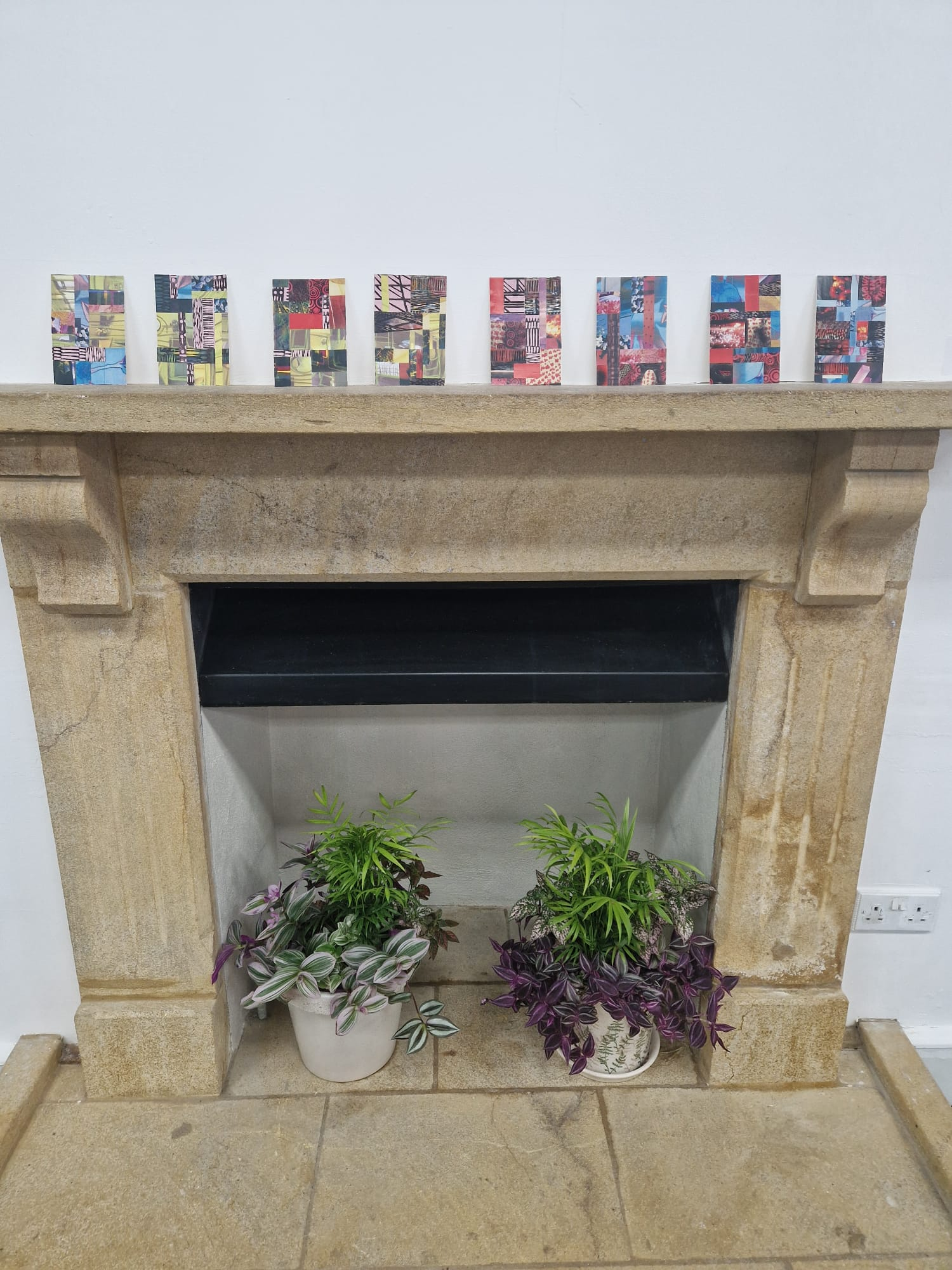 A stone fireplace with a recessed black section below, flanked by two potted plants. Above the fireplace, seven colourful art pieces are arranged in a row, showcasing various abstract designs on a white wall background.
