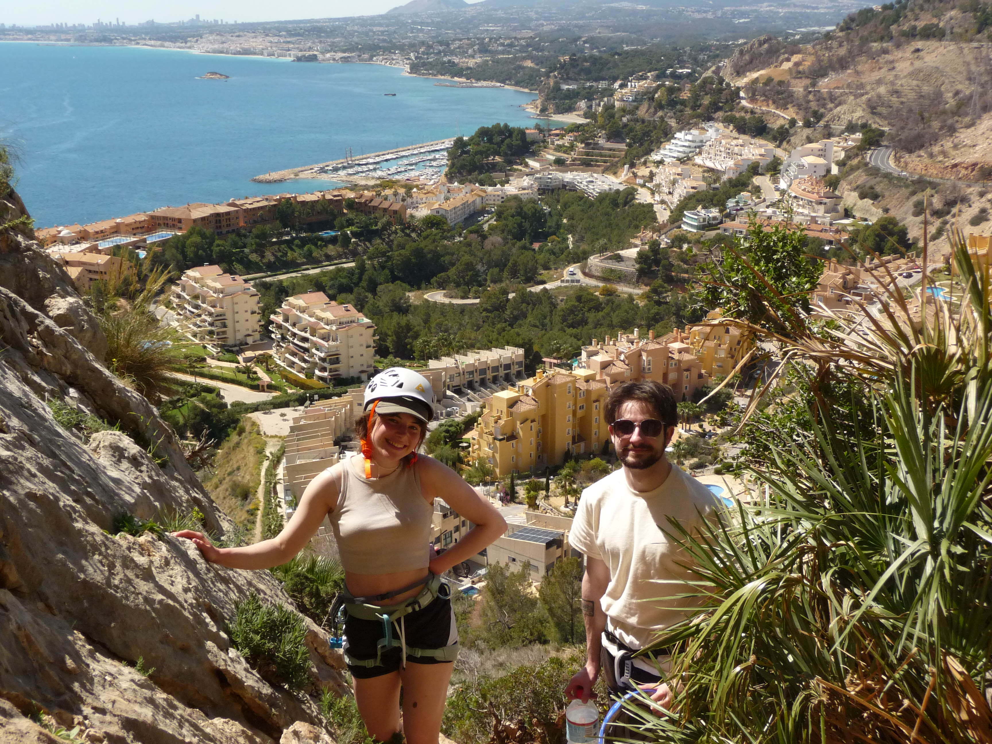 Two hikers on rocky hillside overlooking Mediterranean coastal town with blue sea, white buildings, and marina below.